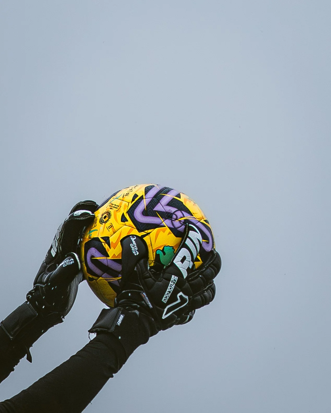A goalkeeper hand holding a colorful football against an overcast sky.