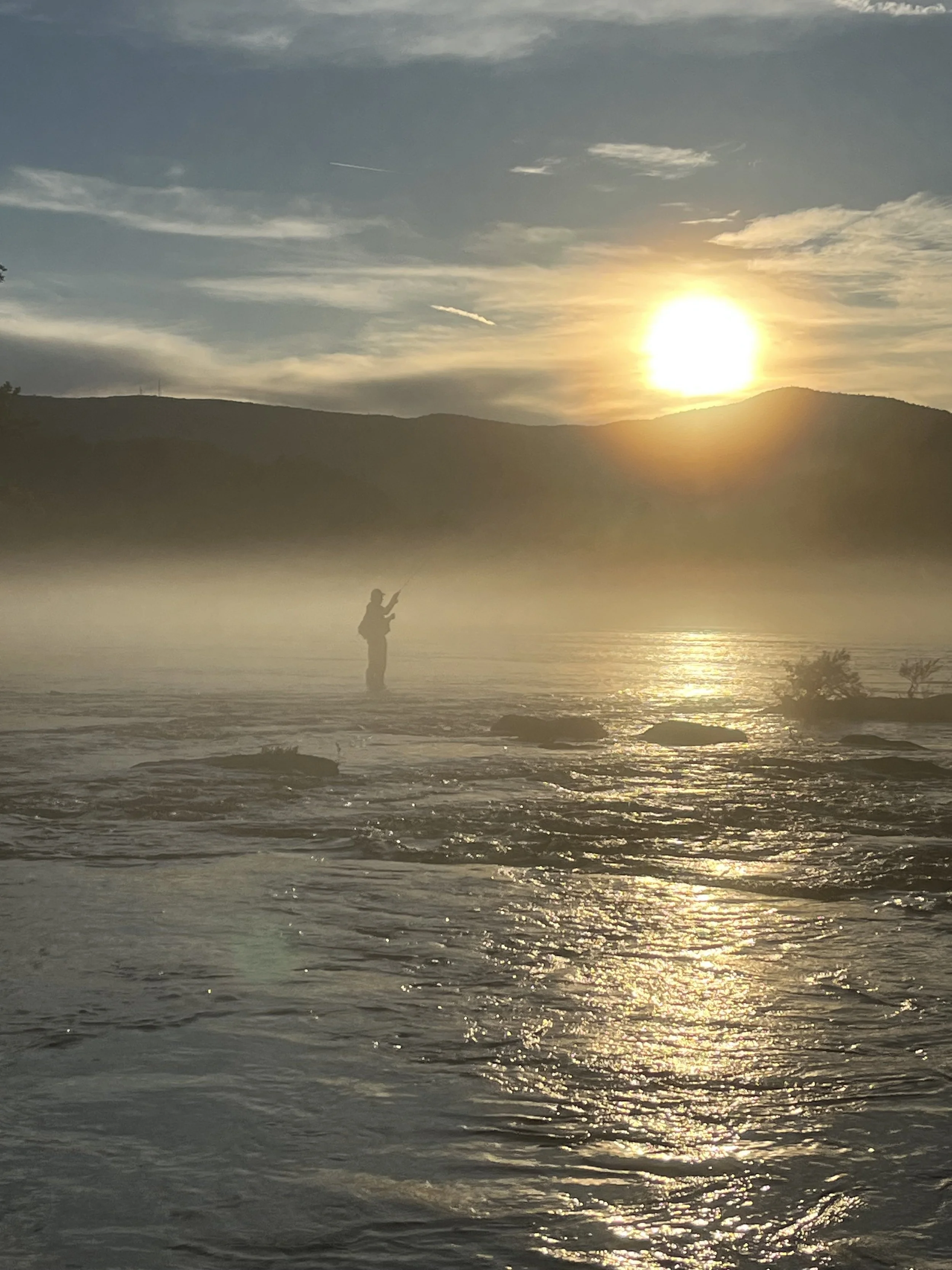 Sunrise over a forested Appalachian ridgeline in East Tennessee, an angler silhouetted while fly fishing the river below.