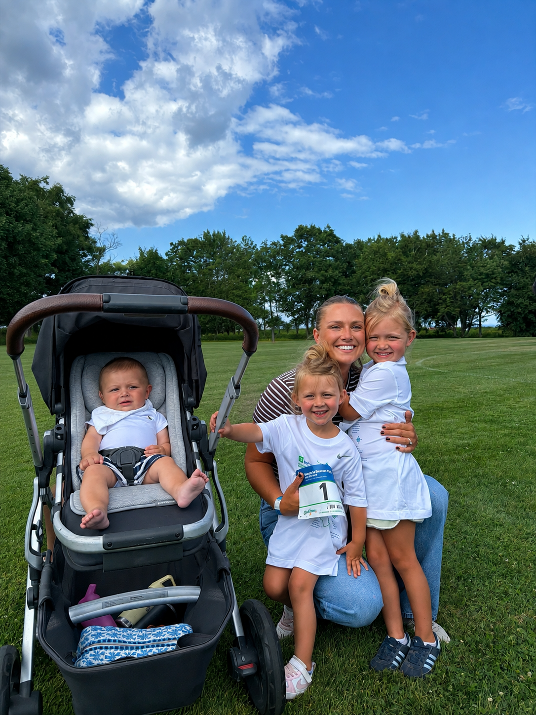 A woman with three young girls smiling and hugging outdoors on a grassy field, with a stroller holding a baby nearby and a background of trees and a partly cloudy sky.