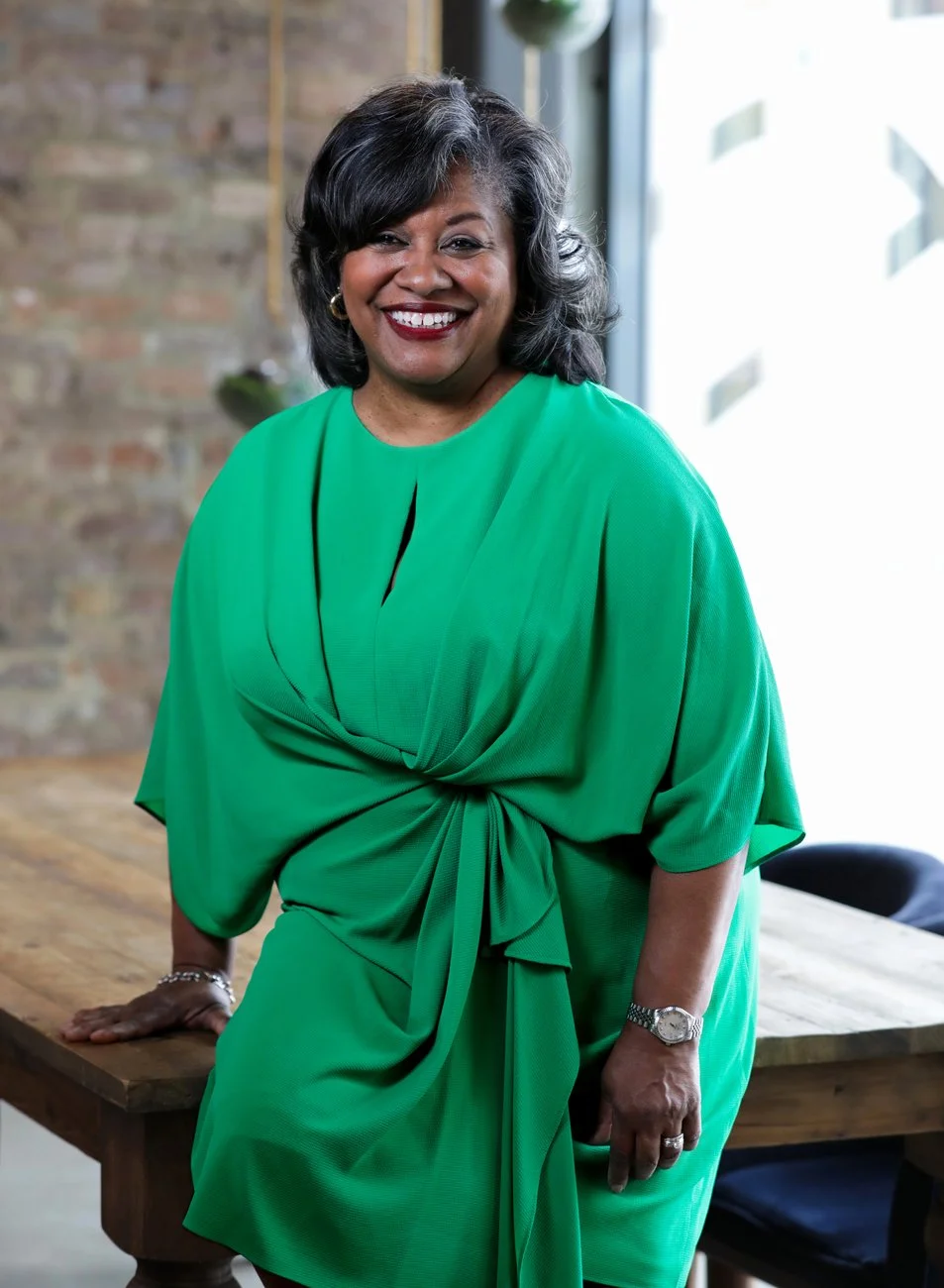 A smiling woman with dark, wavy hair wearing a bright green dress standing in a modern office with a brick wall and large window in the background.