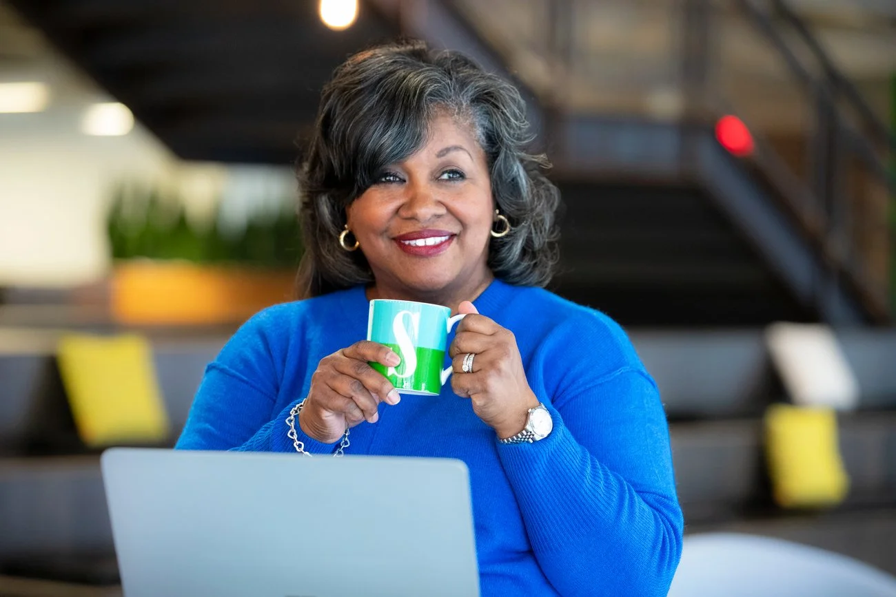 A woman wearing a blue sweater is smiling and holding a green and white coffee mug in a modern indoor setting.