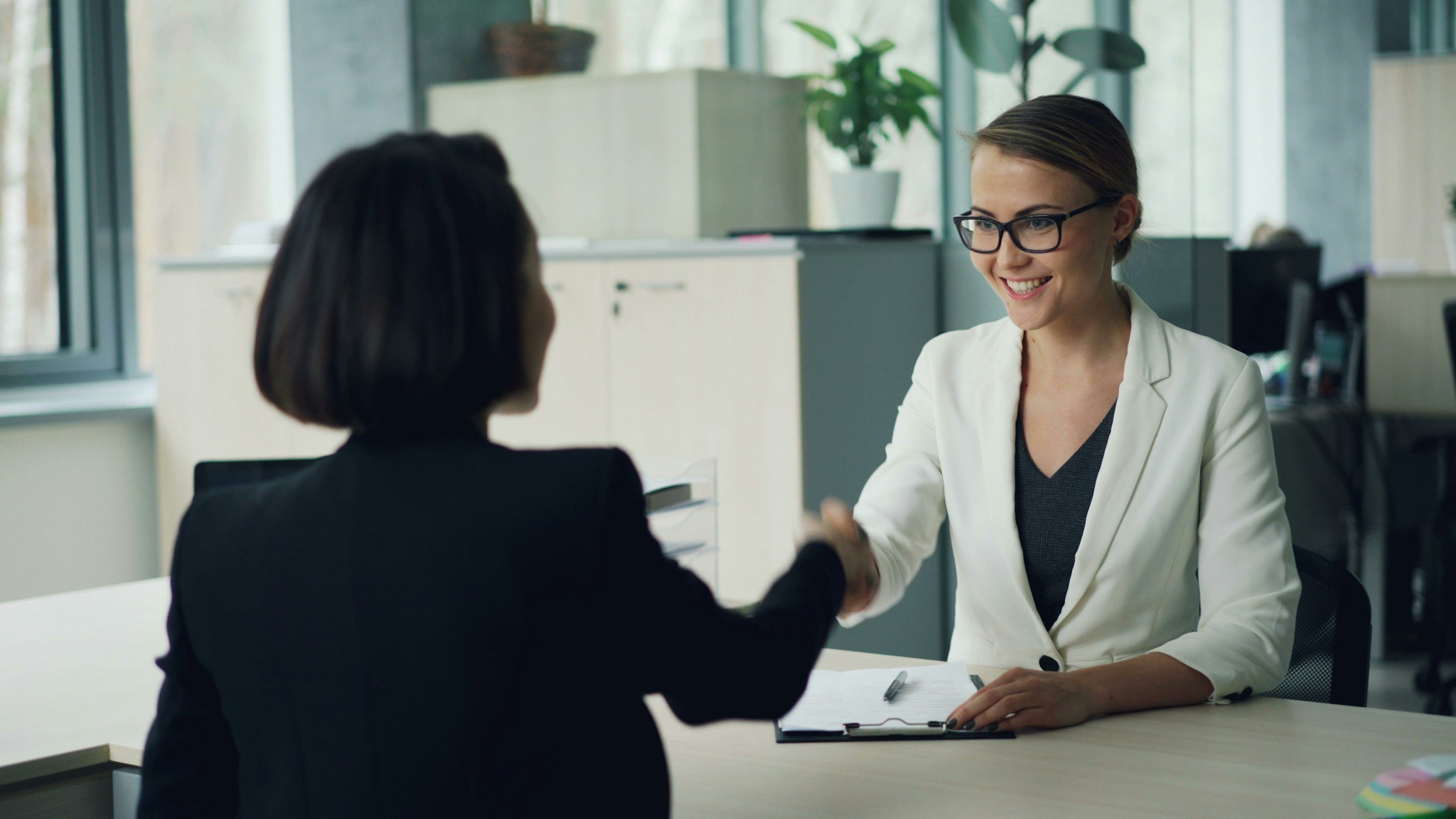 Two women shaking hands in an office setting, one seated and smiling, the other facing away.