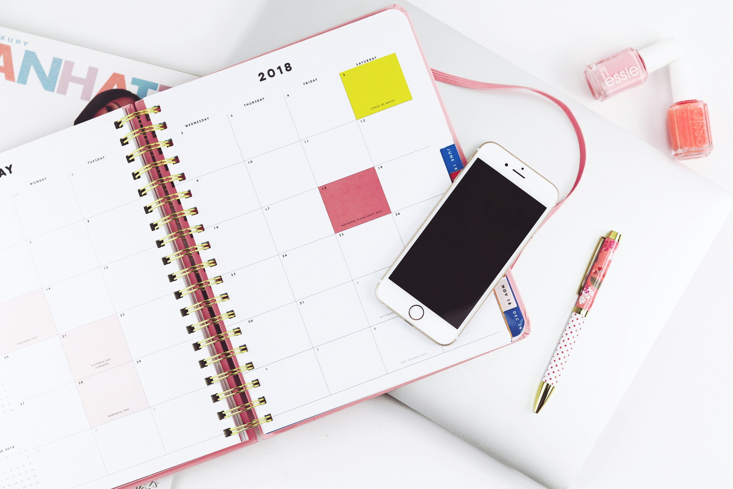 Open 2018 planner with weekly layout, yellow and red sticky notes, a white iPhone with black screen, pink nail polish bottles, floral-patterned pen, and a pink-tipped pen on a white surface.