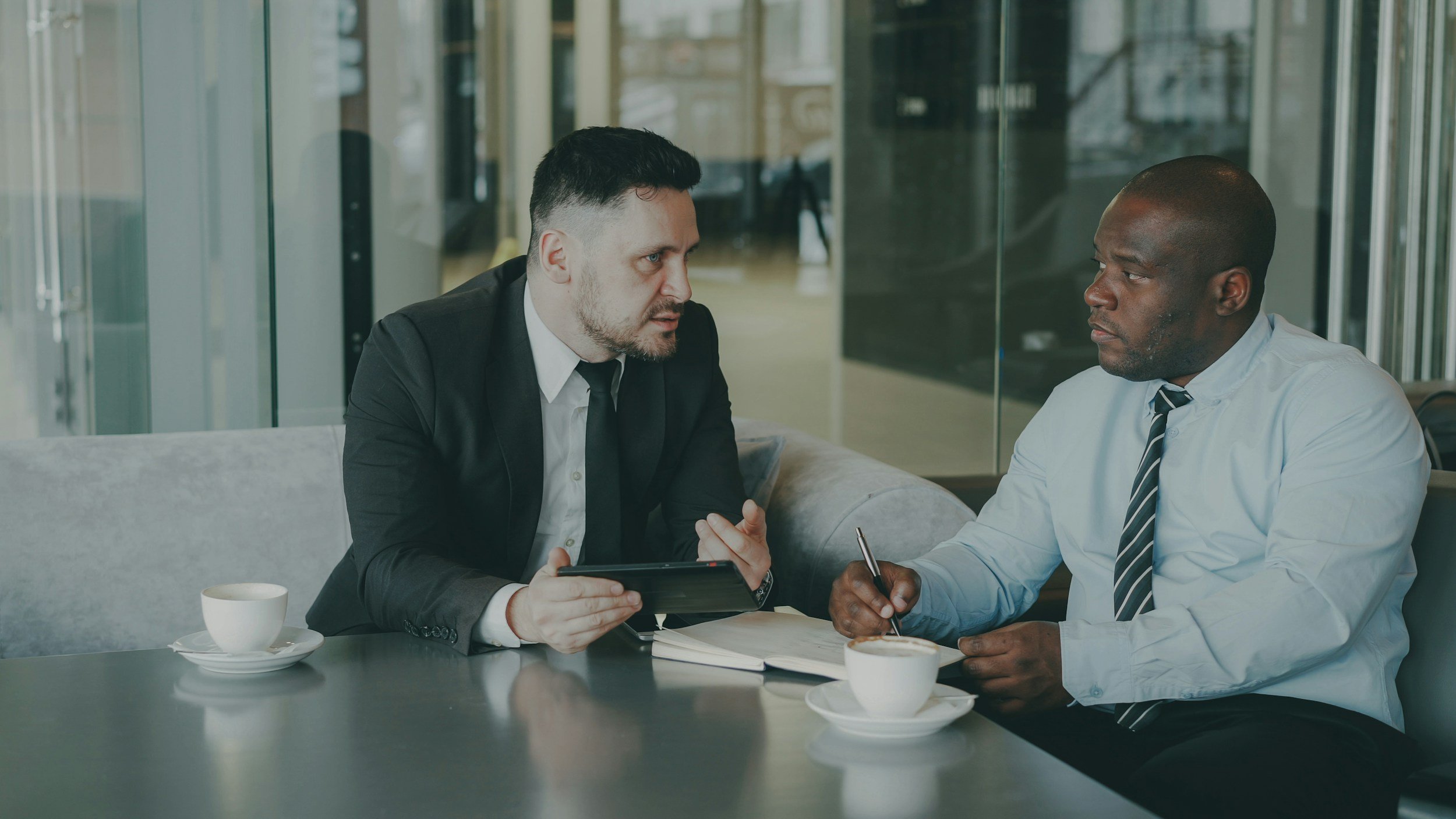 Two men in business attire having a serious conversation at a table in a modern office. One man is holding a tablet, and both have pens and notebooks in front of them. There are two cups of coffee on the table.