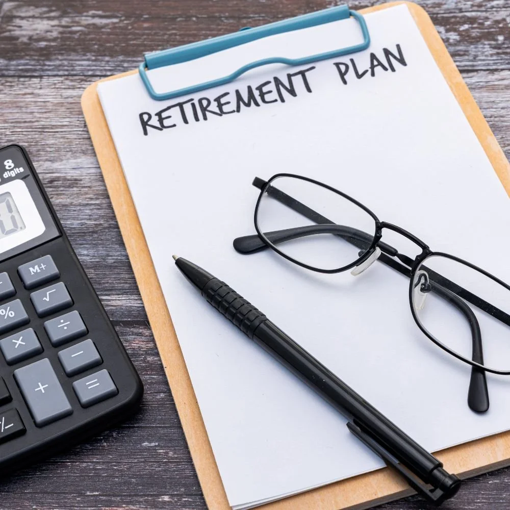 A clipboard with 'Retirement Plan' written on it, eyeglasses, a black pen, and a calculator on a wooden desk.