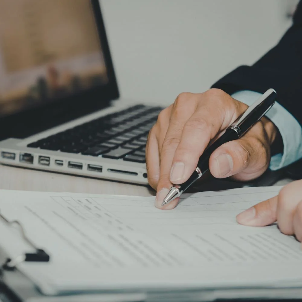 A person in a black suit holding a pen and writing on paper at a desk with an open laptop in the background