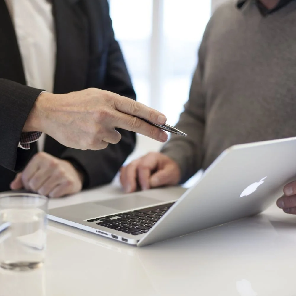 Two people discussing something over a laptop and a tablet, with a glass of water on the table.