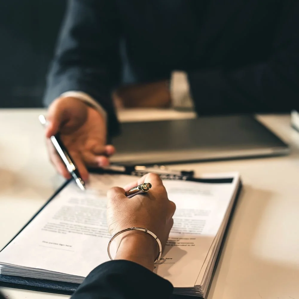 Person signing a document at a desk with a pen, with a laptop and other papers nearby.