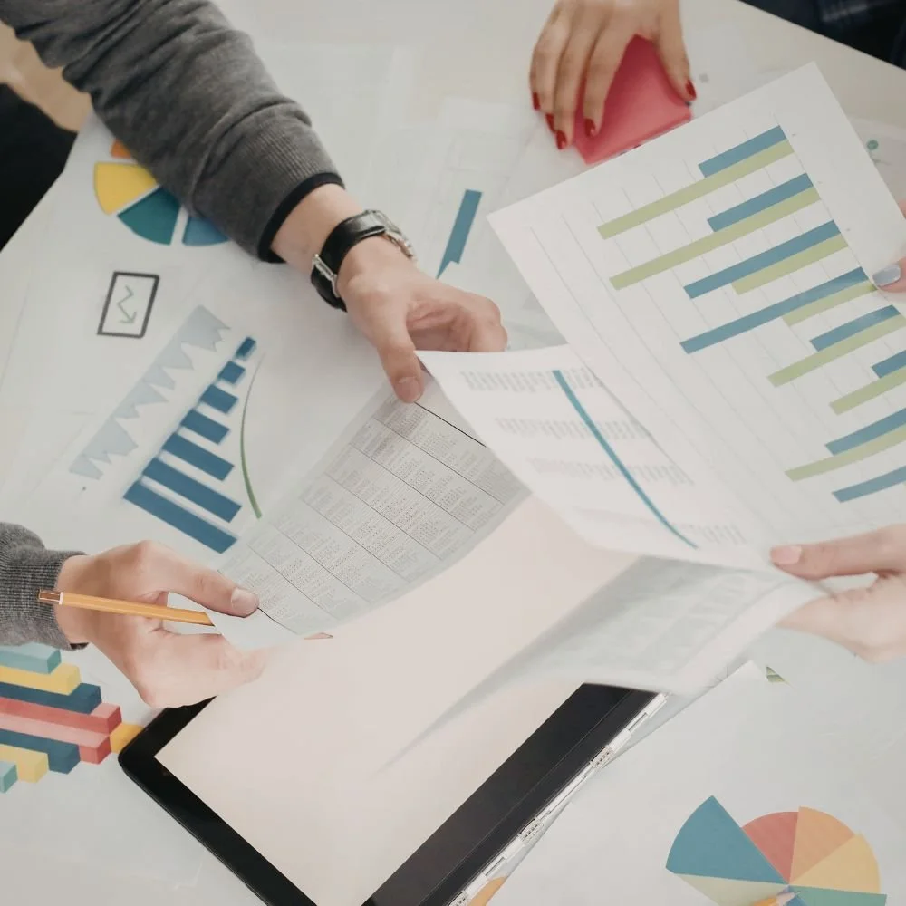 People discussing and analyzing printed charts and data sheets on a white table, with colorful graphs and a laptop in the background.