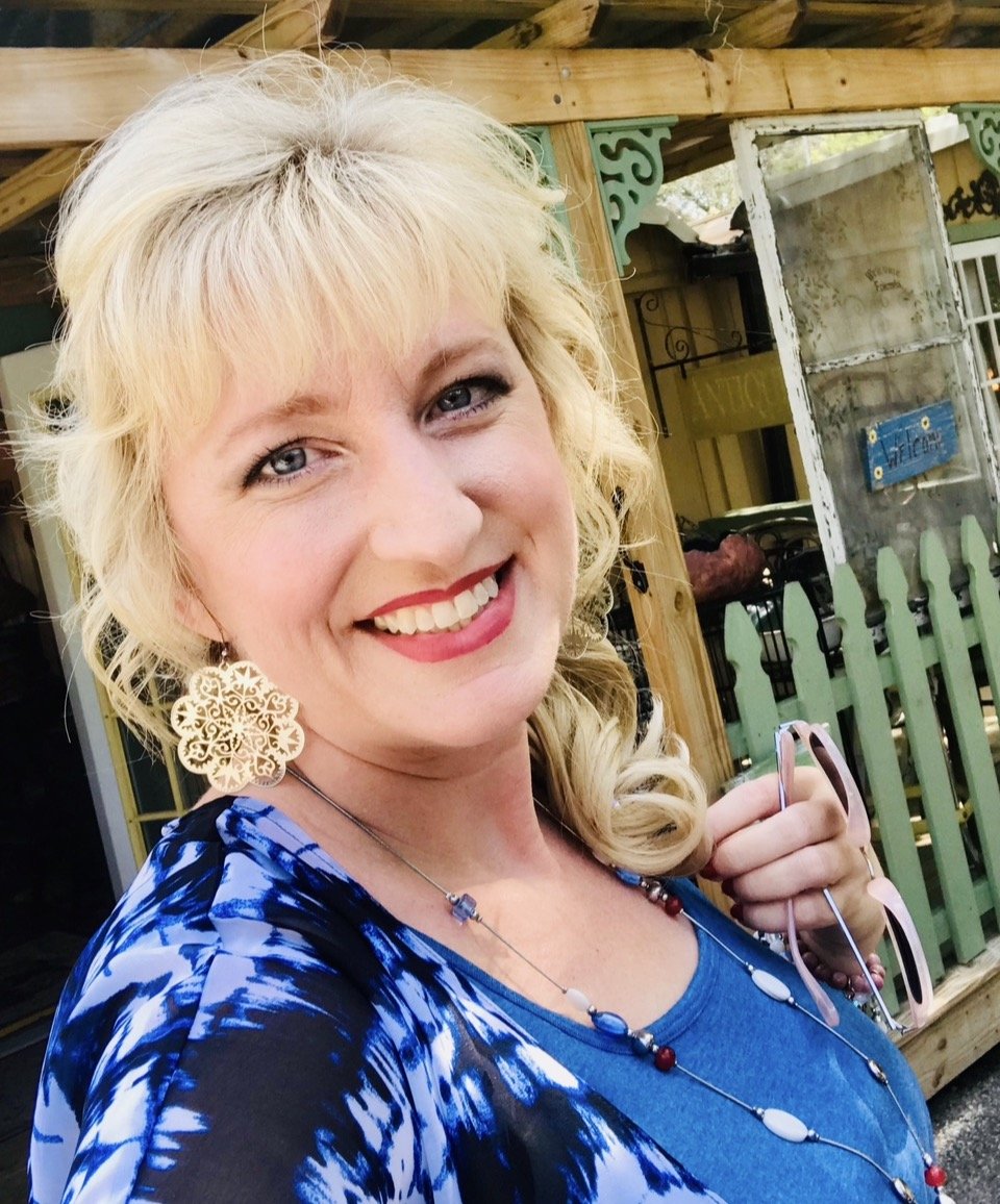 A woman with blonde hair smiling, wearing large earrings and a blue patterned top, holding pink sunglasses outdoors with a wooden fence and a building in the background.