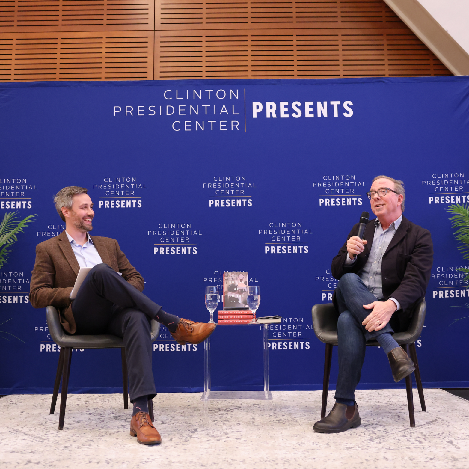 Two men are seated on stage at a book event. The man on the left is wearing a brown blazer, white shirt, dark pants, and brown shoes. The man on the right is wearing a black blazer, light shirt, jeans, and boots, and is holding a microphone. Behind them is a blue backdrop with the text "Clinton Presidential Center Presents" repeated. In front of them is a small table with copies of a book titled "House of Smoke" and two glasses of water.