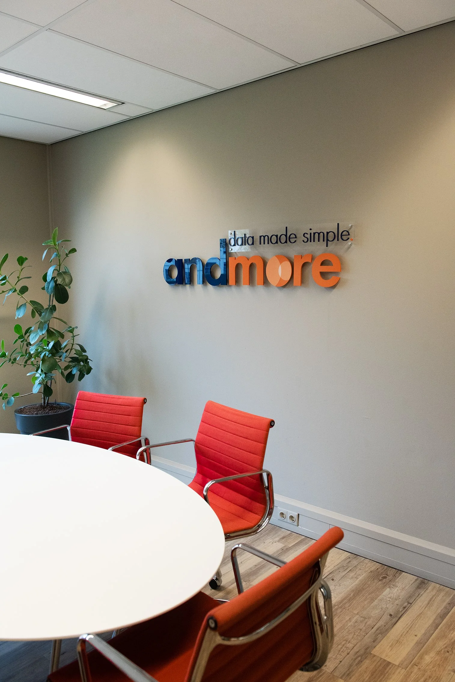 Een vergaderruimte met een wit rond bureau en rode vergaderstoelen, een plant links en een muur met het logo 'andmore' en de slogan 'data made simple'.