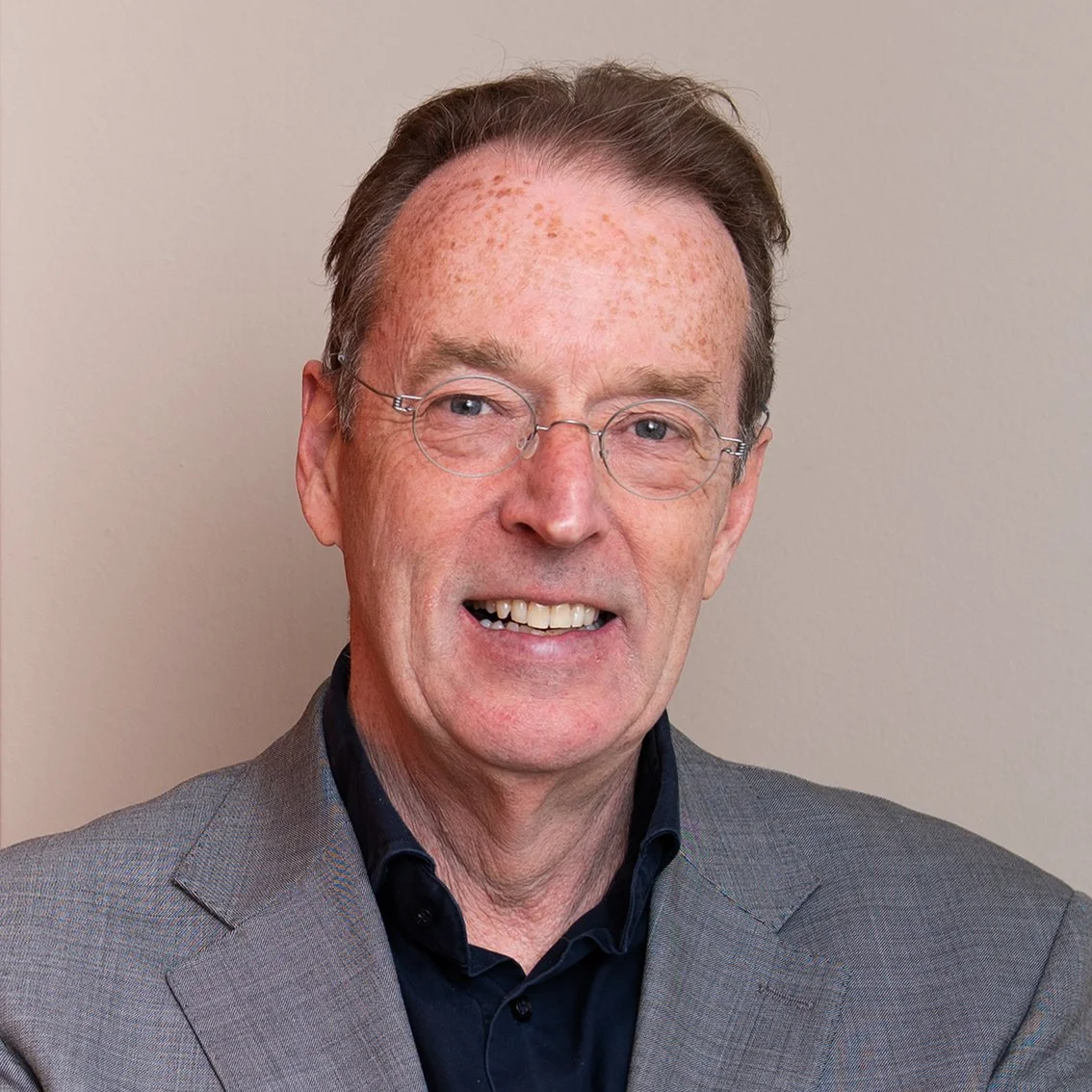 Portret van een man met bril, grijs pak, en een glimlach, staande voor een lichte achtergrond.