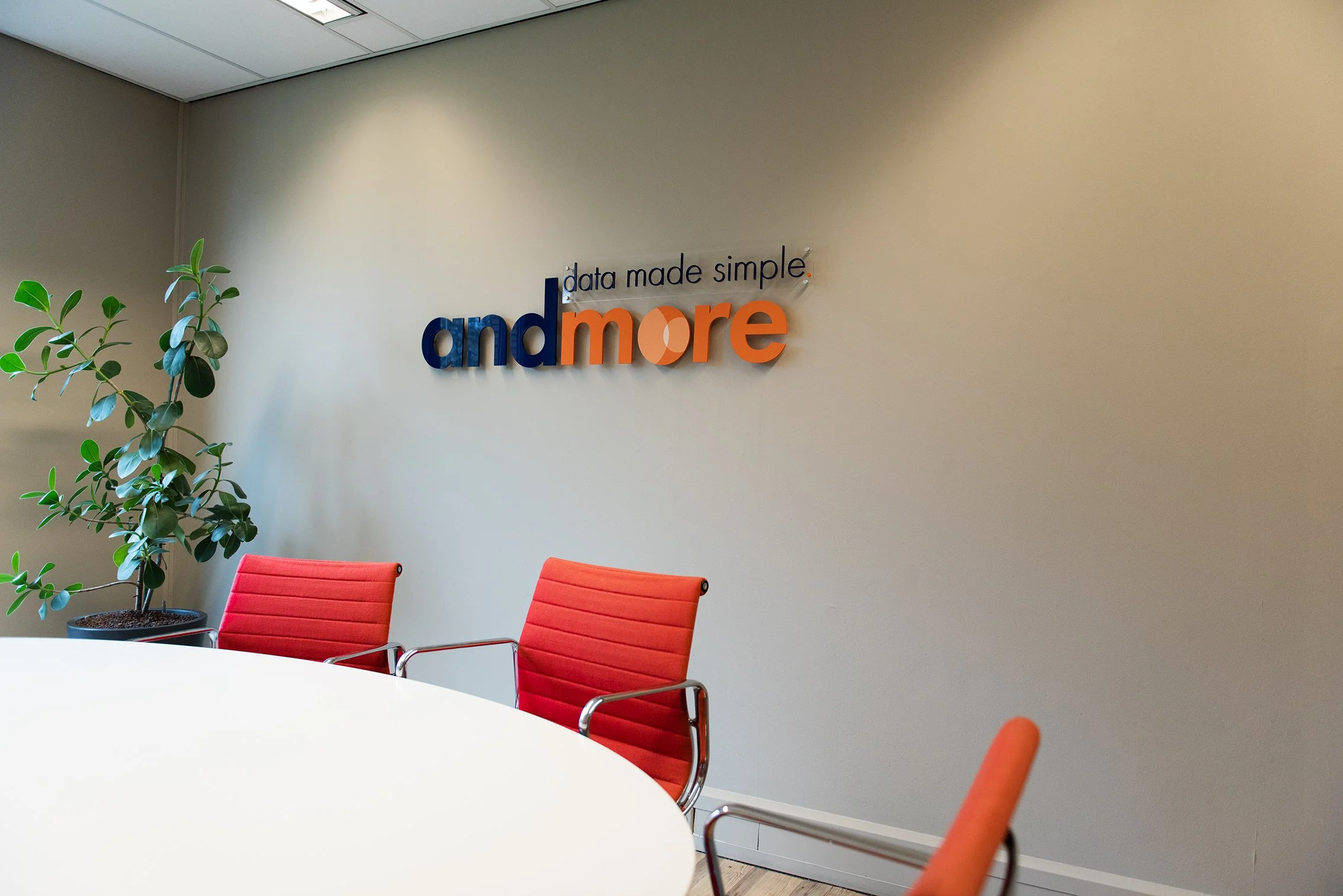 Een vergaderruimte met een witte tafel, rood/oranje stoelen, een groene plant en een muur met het logo 'andmore' en de slogan 'data made simple'.
