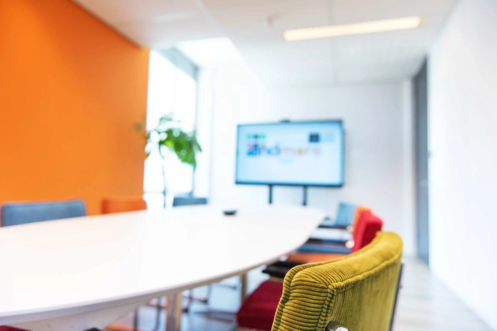 Een vergaderruimte met een wit tafel, kleurrijke stoelen en een grote digitale scherm aan de muur.