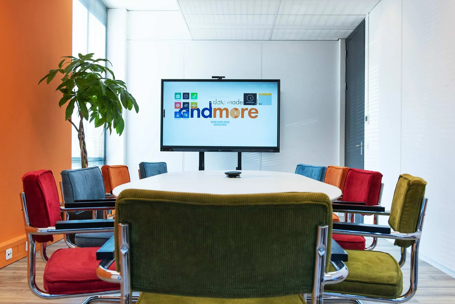Vergaderruimte met kleurrijke stoelen, een grote tafel, een grote scherm met logo, planten en witte en oranje muren.