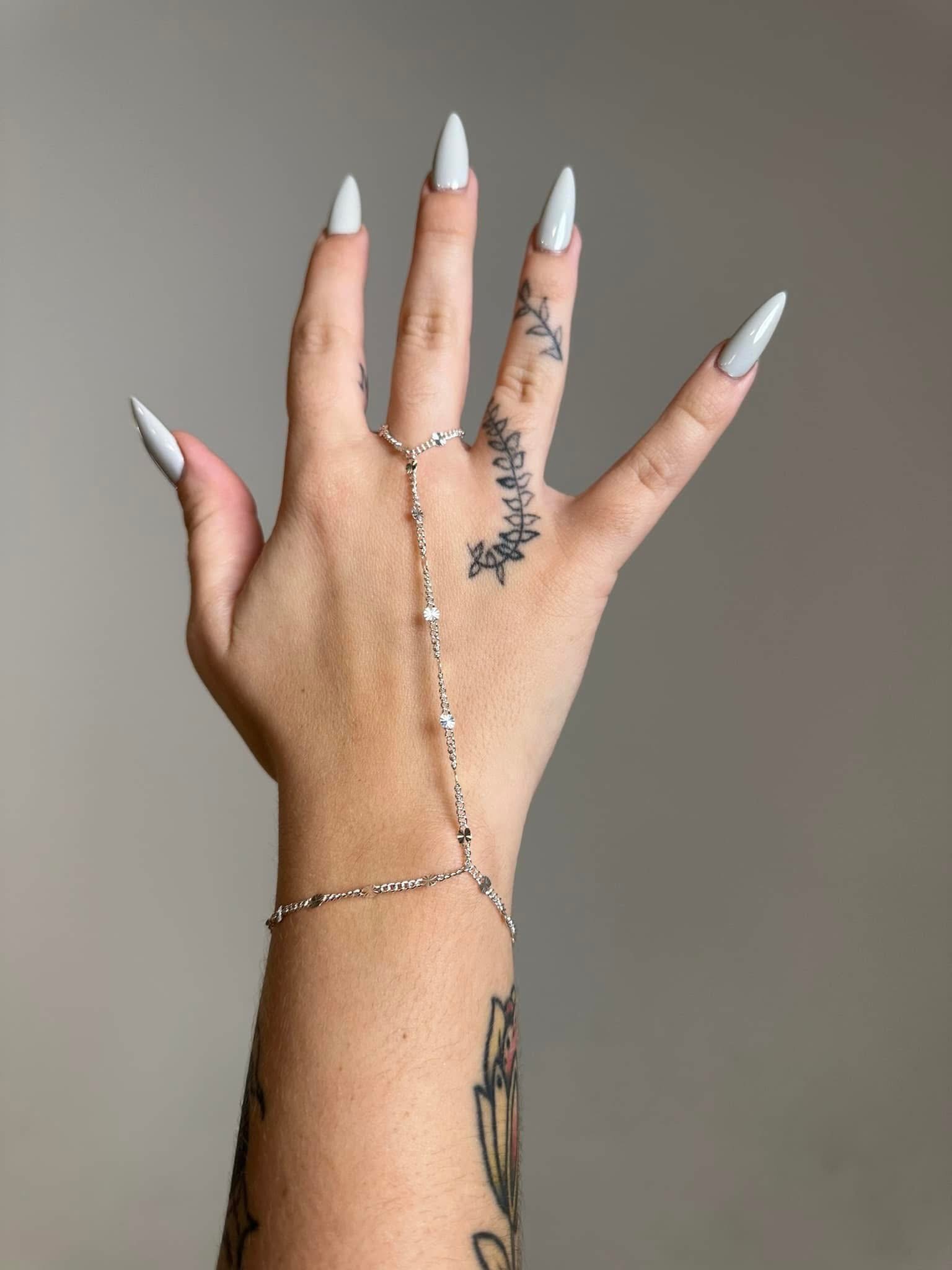A hand with long, white, pointed nails, multiple rings, a delicate chain bracelet, and tattoos on the fingers and forearm against a plain background.