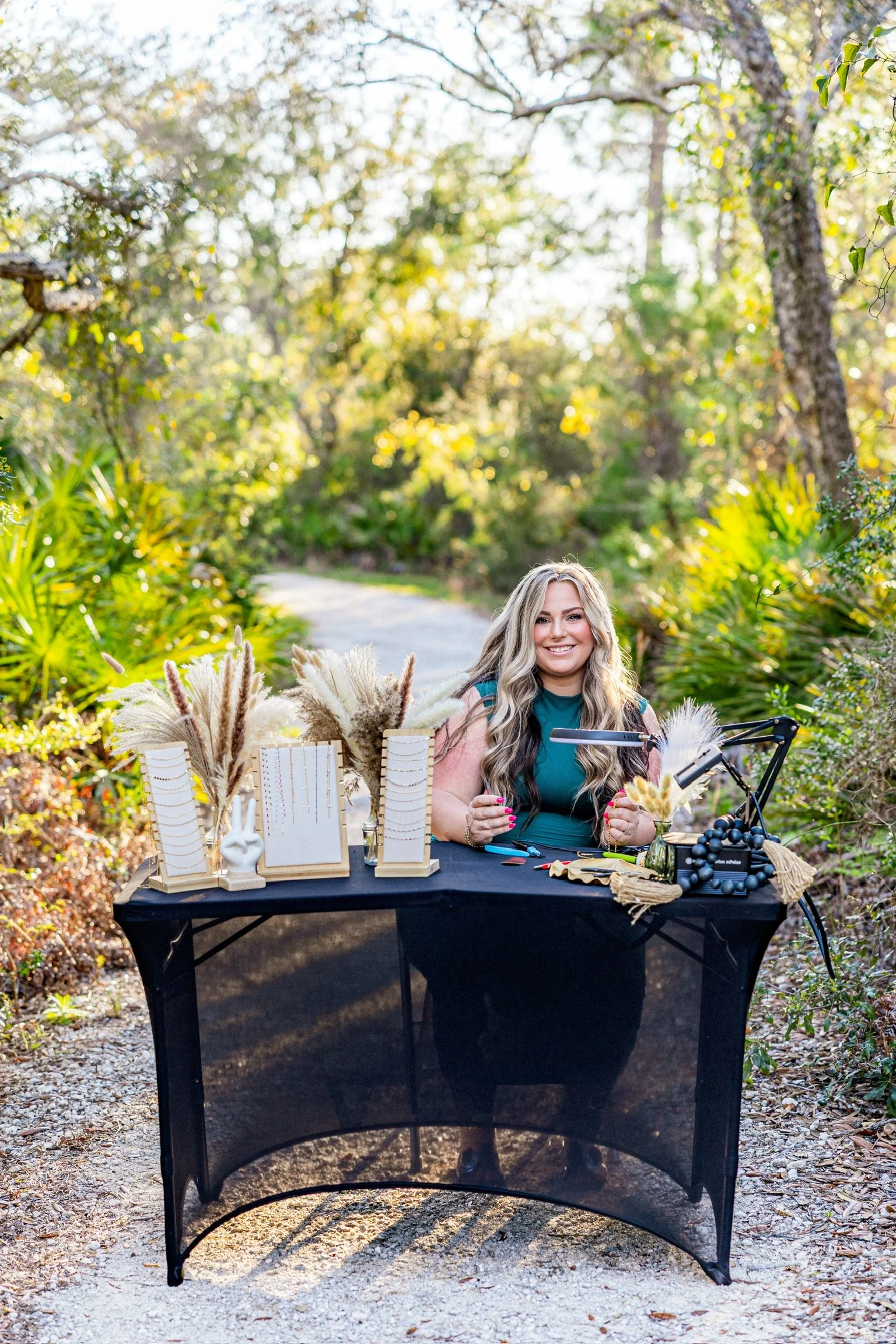 A woman with long blonde hair sitting at a table outdoors in a natural setting with trees and greenery. The table is decorated with various items including display stands with jewelry, decorative grasses, and other craft materials.
