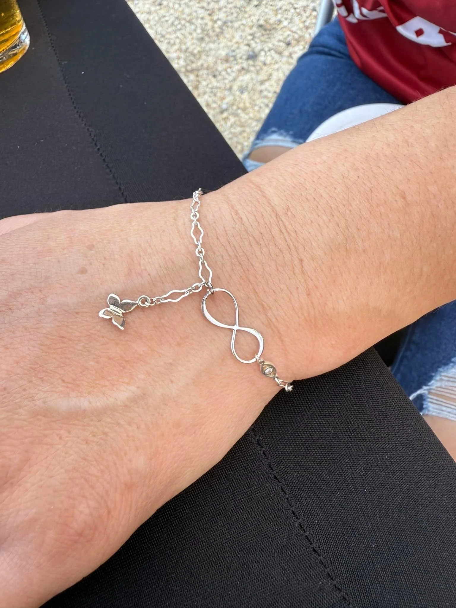 A person's arm resting on a black table, wearing a silver bracelet with a butterfly charm and an infinity symbol charm.