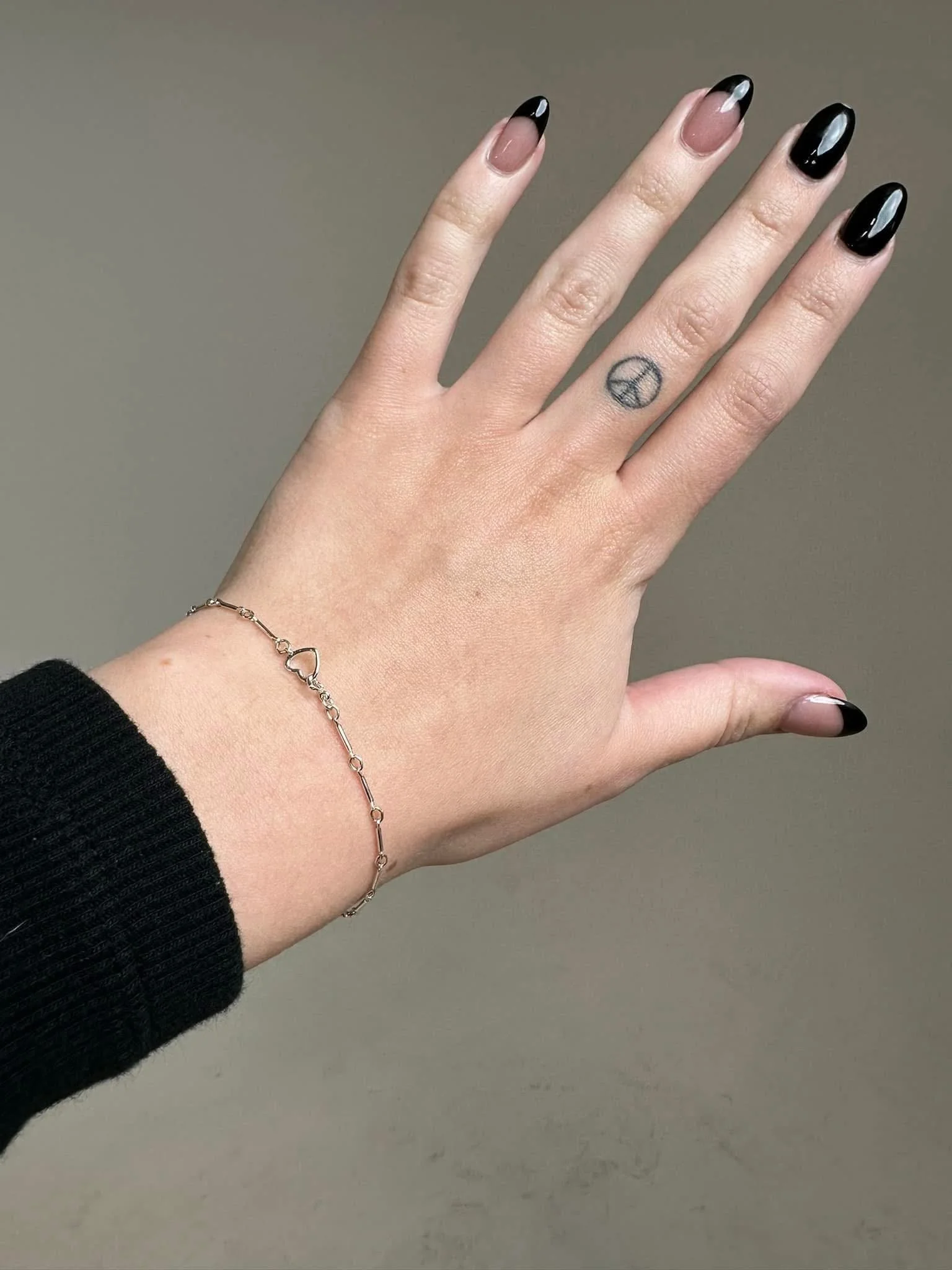 A person's hand with black-tipped acrylic nails, a peace sign tattoo on the middle finger, a silver chain bracelet with heart and key charms, and a black long-sleeve shirt or sweater.