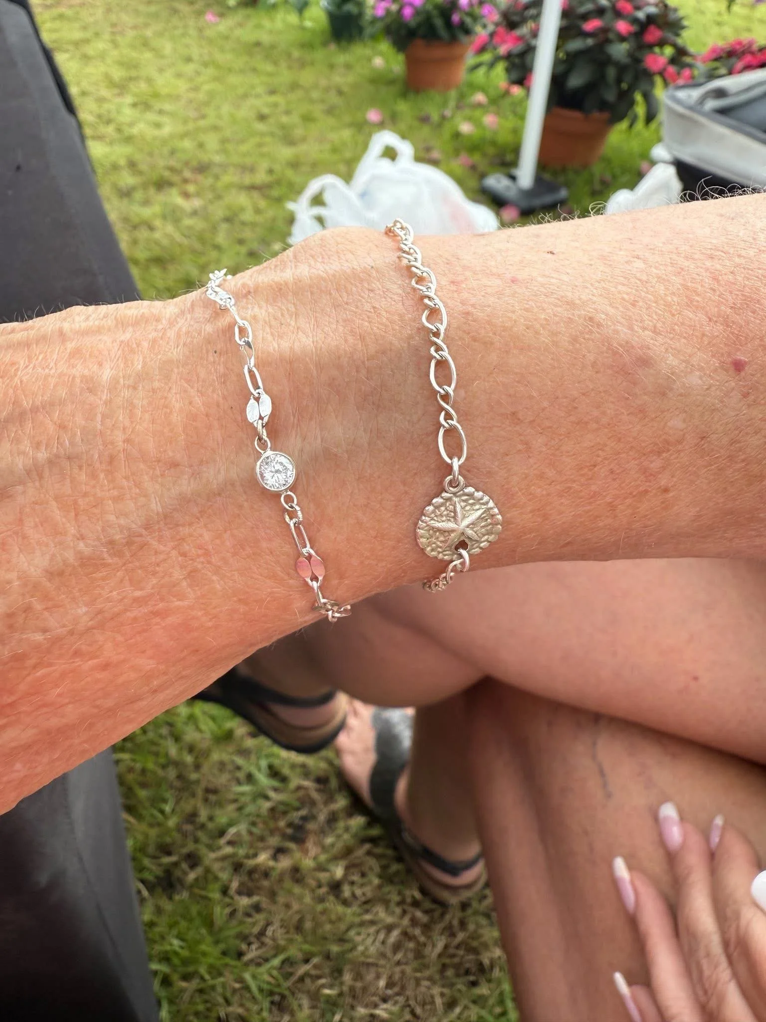 Close-up of a person's wrist with silver bracelets, one with a diamond charm, with a hand resting on a person's knee outdoors, with potted plants and grass in the background.