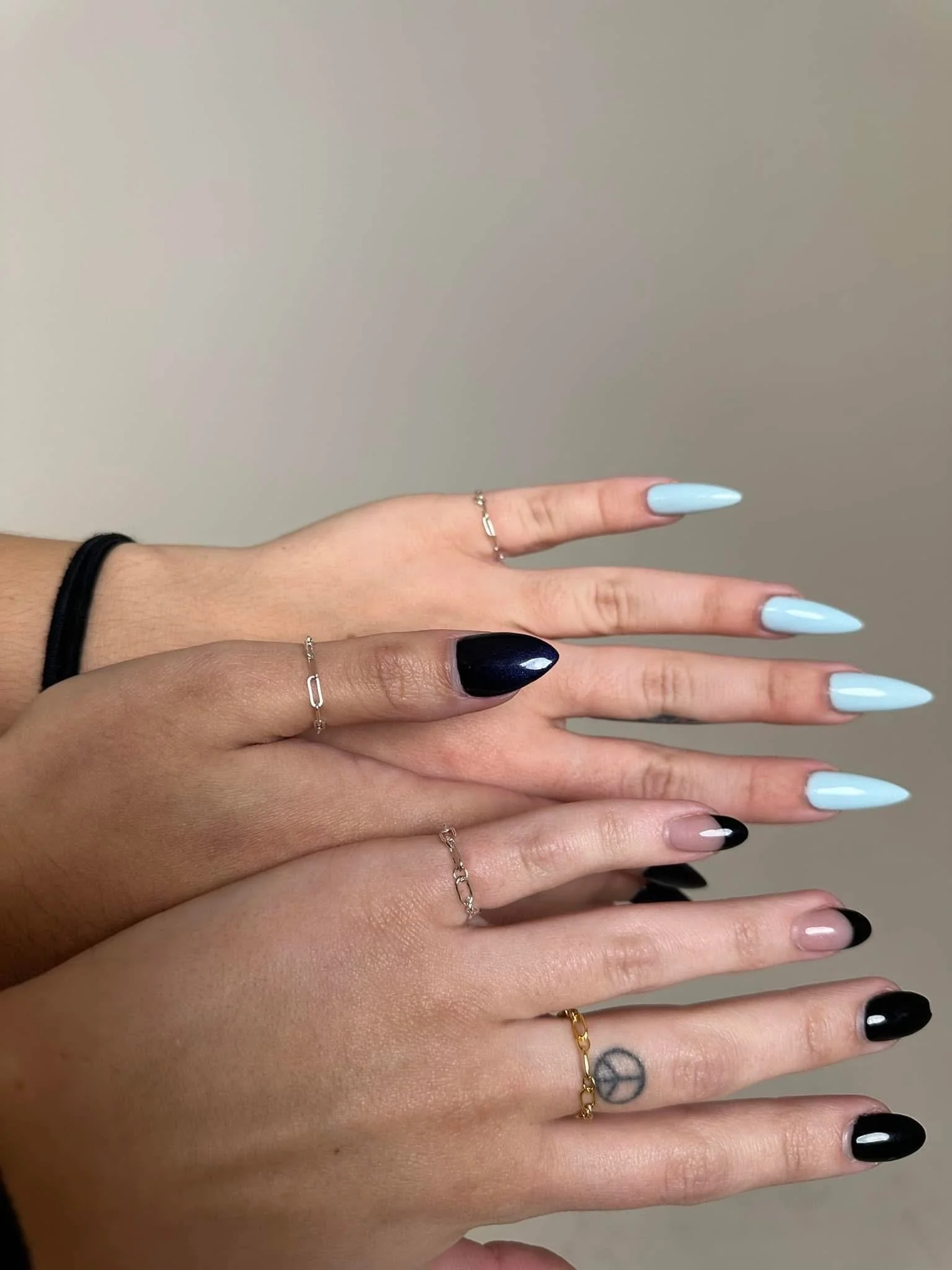 Multiple hands with manicured nails, some painted light blue, black, and pink, wearing delicate rings and bracelets, with a tattoo of a peace symbol on the wrist.