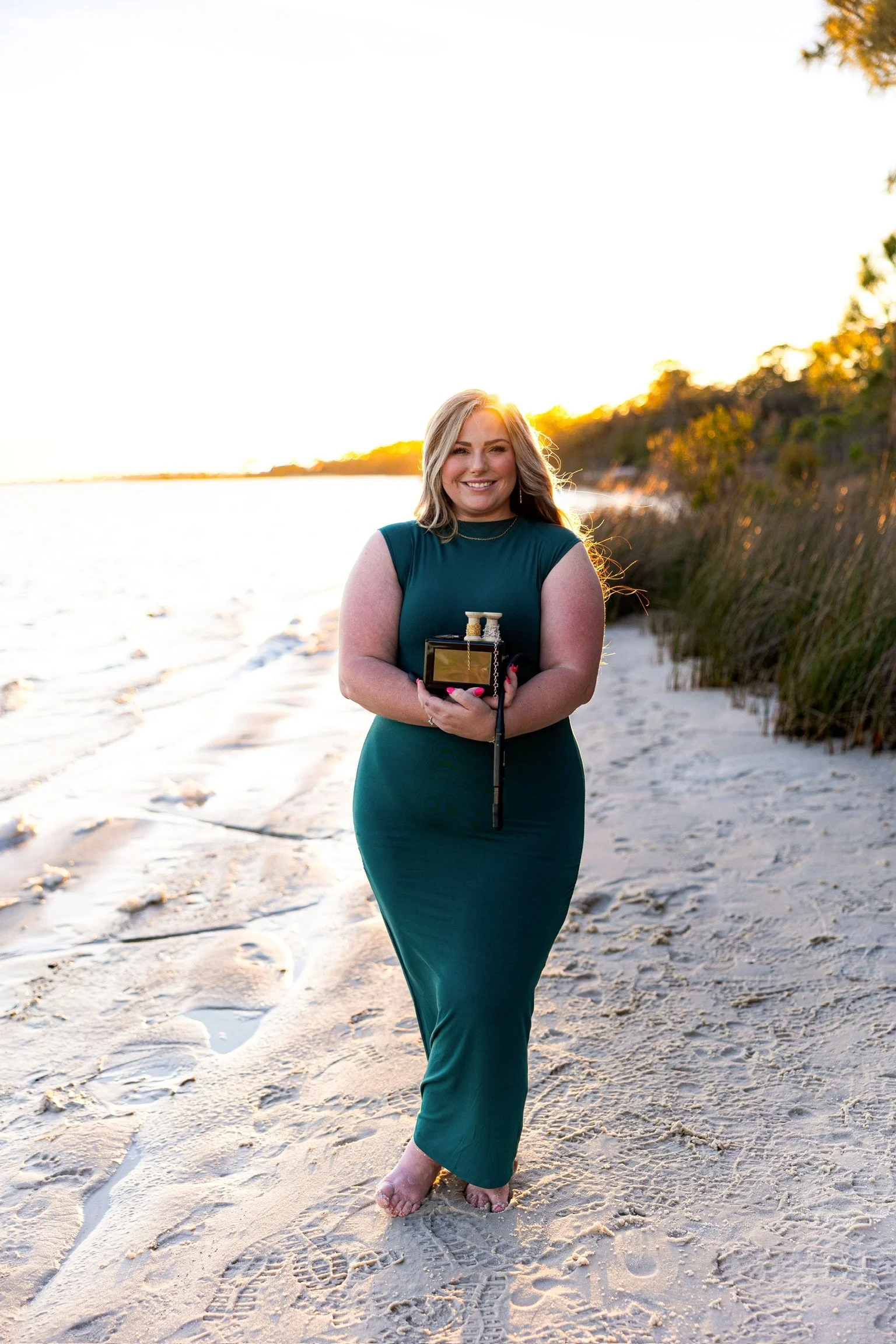 A woman in a green dress standing barefoot on the beach during sunset, holding a small black box with sewing spools inside, smiling at the camera.
