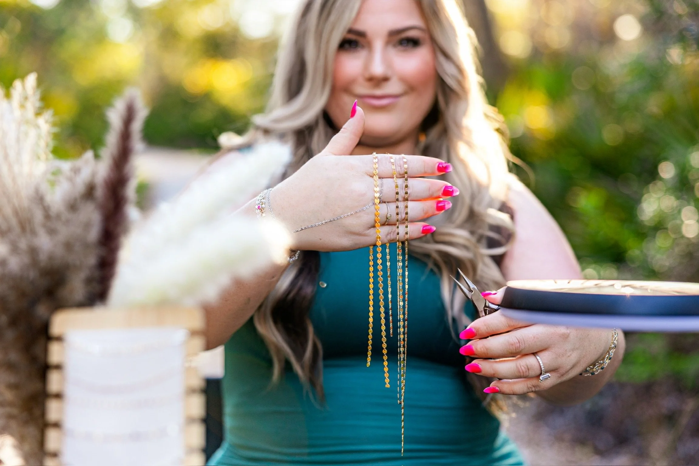 A woman with long blonde hair and pink nail polish shows her rings and jewelry while sitting outdoors, holding a smartphone, with a blurred natural background.