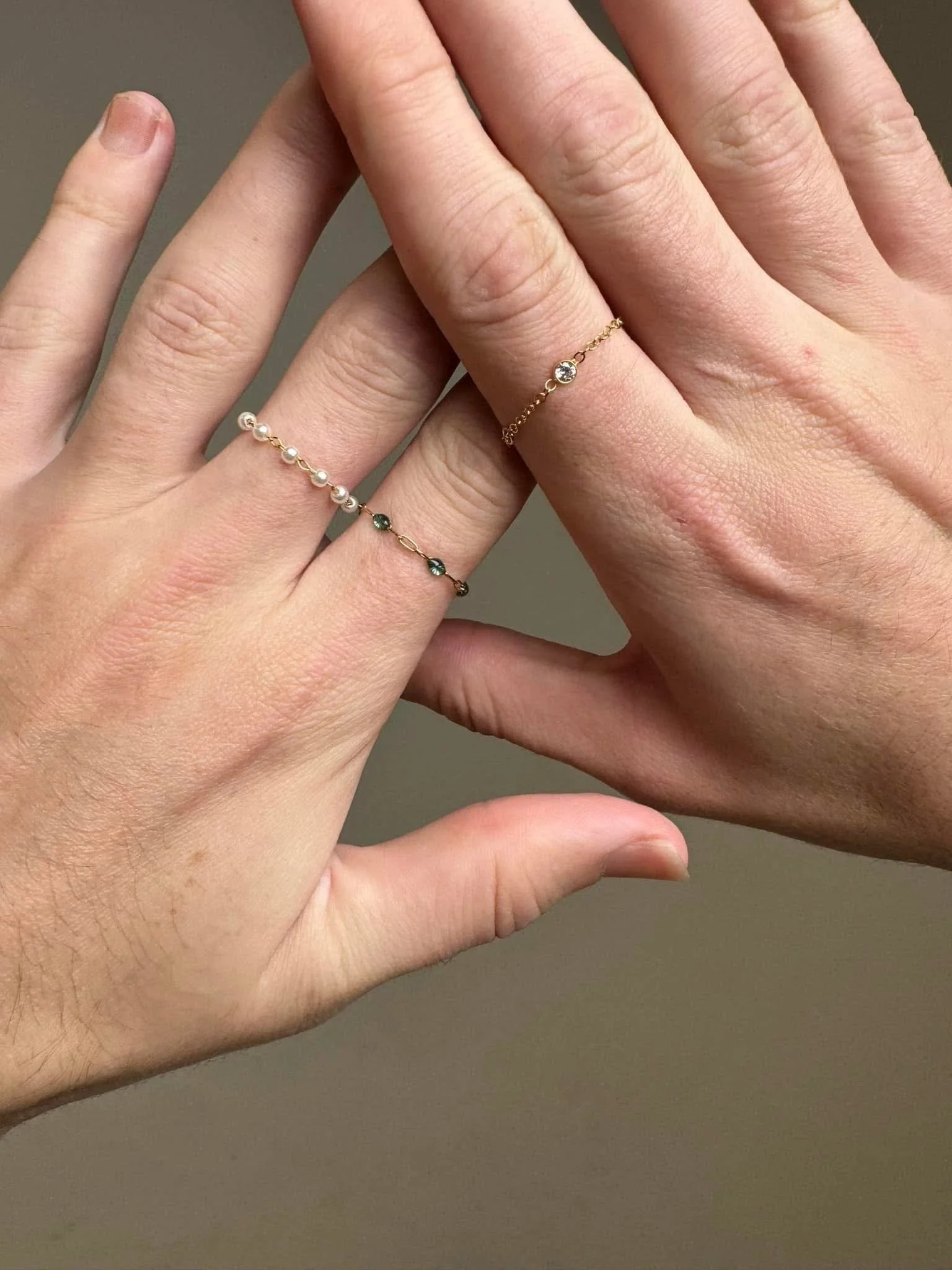 A hand wearing three delicate rings on the fingers, with a plain beige background.