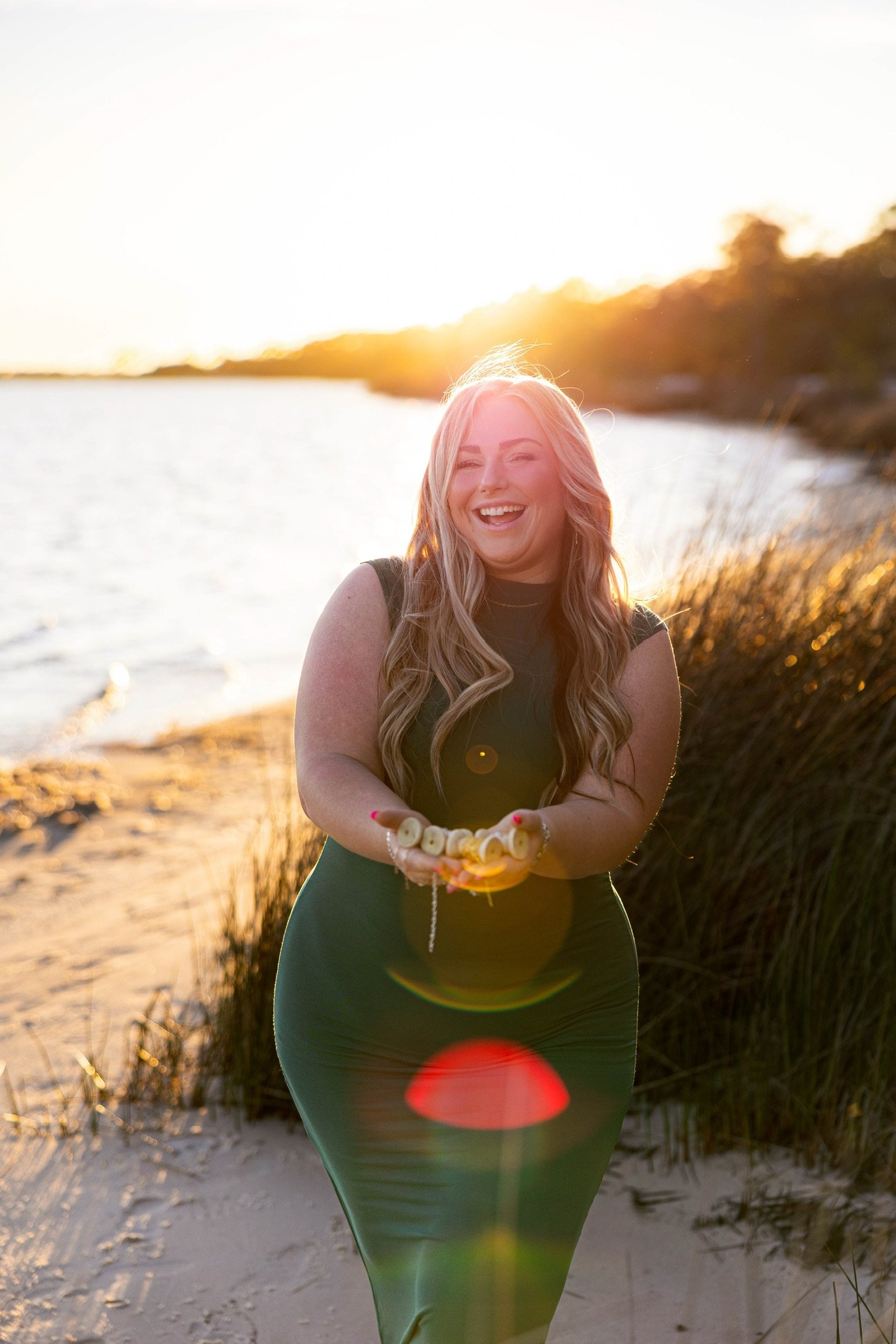 A woman with long, wavy blonde hair smiling and holding a bracelet, standing outdoors at sunset on a beach with sand and dunes, with trees in the background.