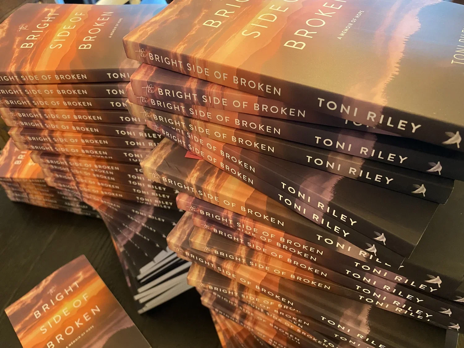 Multiple copies of a book titled 'The Bright Side of Broken' by Toni Riley, stacked in a pile with a sunset or sunrise background on the cover.