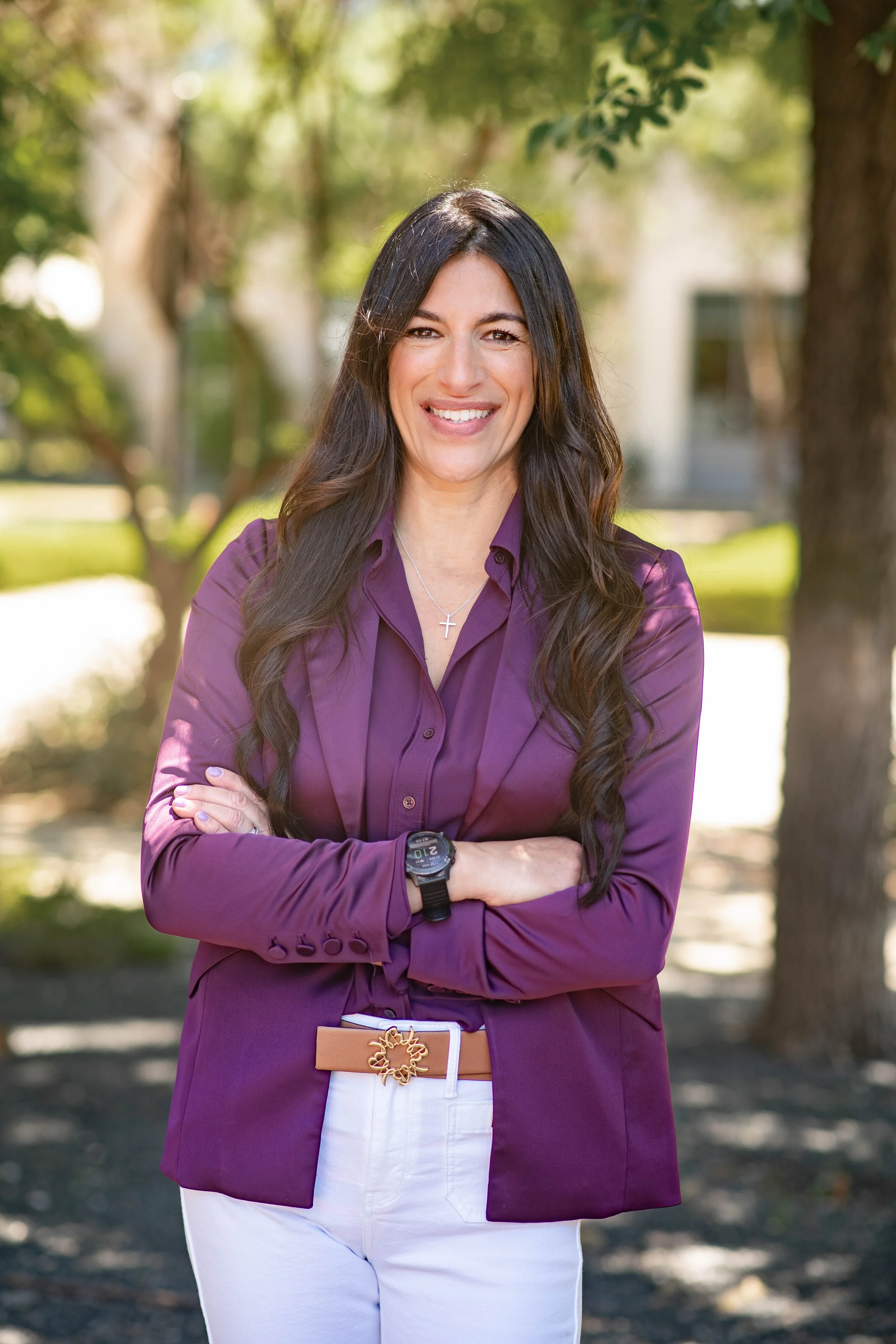 A smiling woman with long dark hair wearing a purple blazer, white pants, a cross necklace, and a watch, standing outdoors with her arms crossed, in a park with trees in the background.