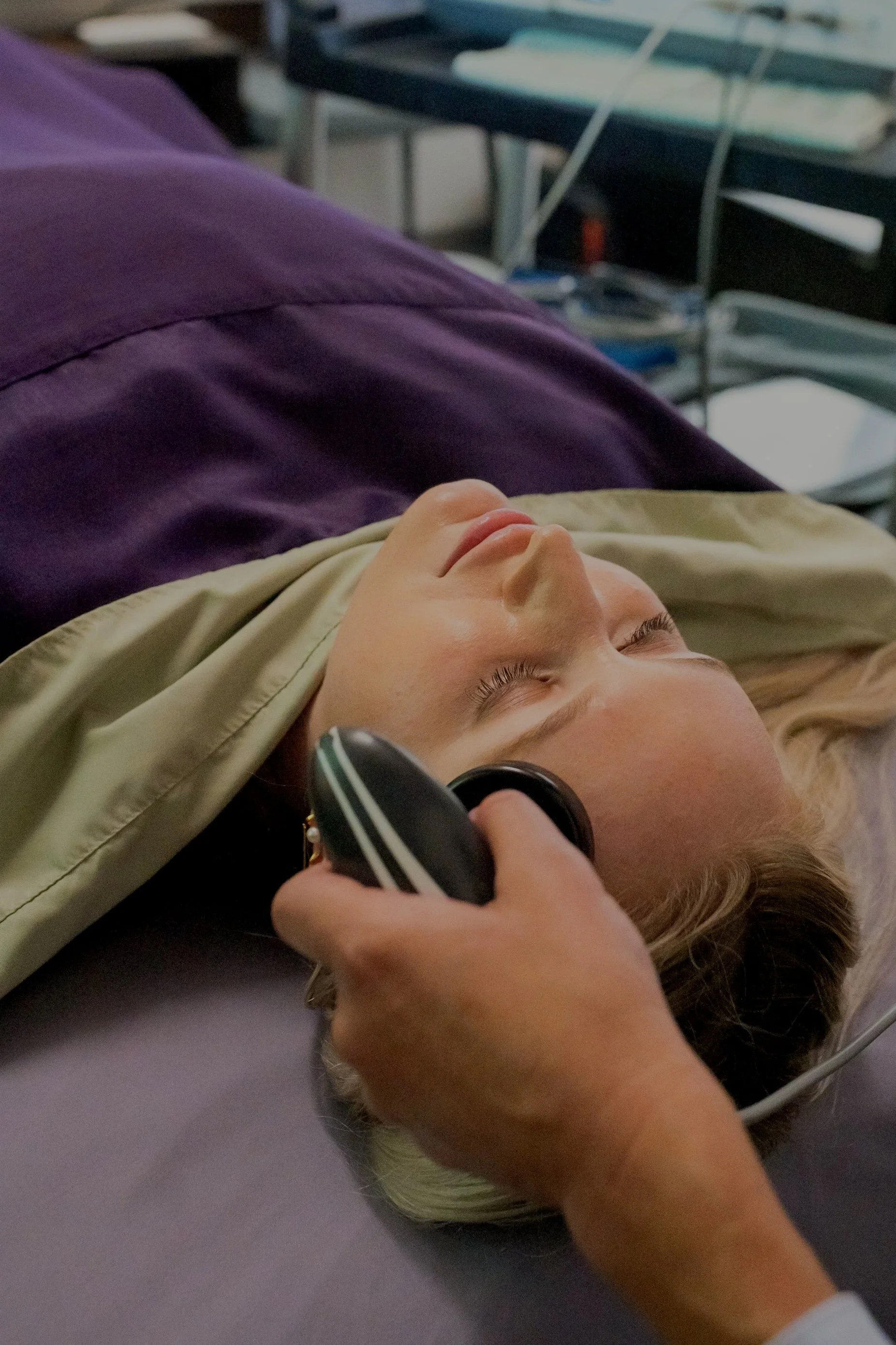 A person lying on a hospital bed with eyes closed, holding a stethoscope to their head, wearing a beige shirt and purple pants, with medical equipment and a hospital bed in the background.