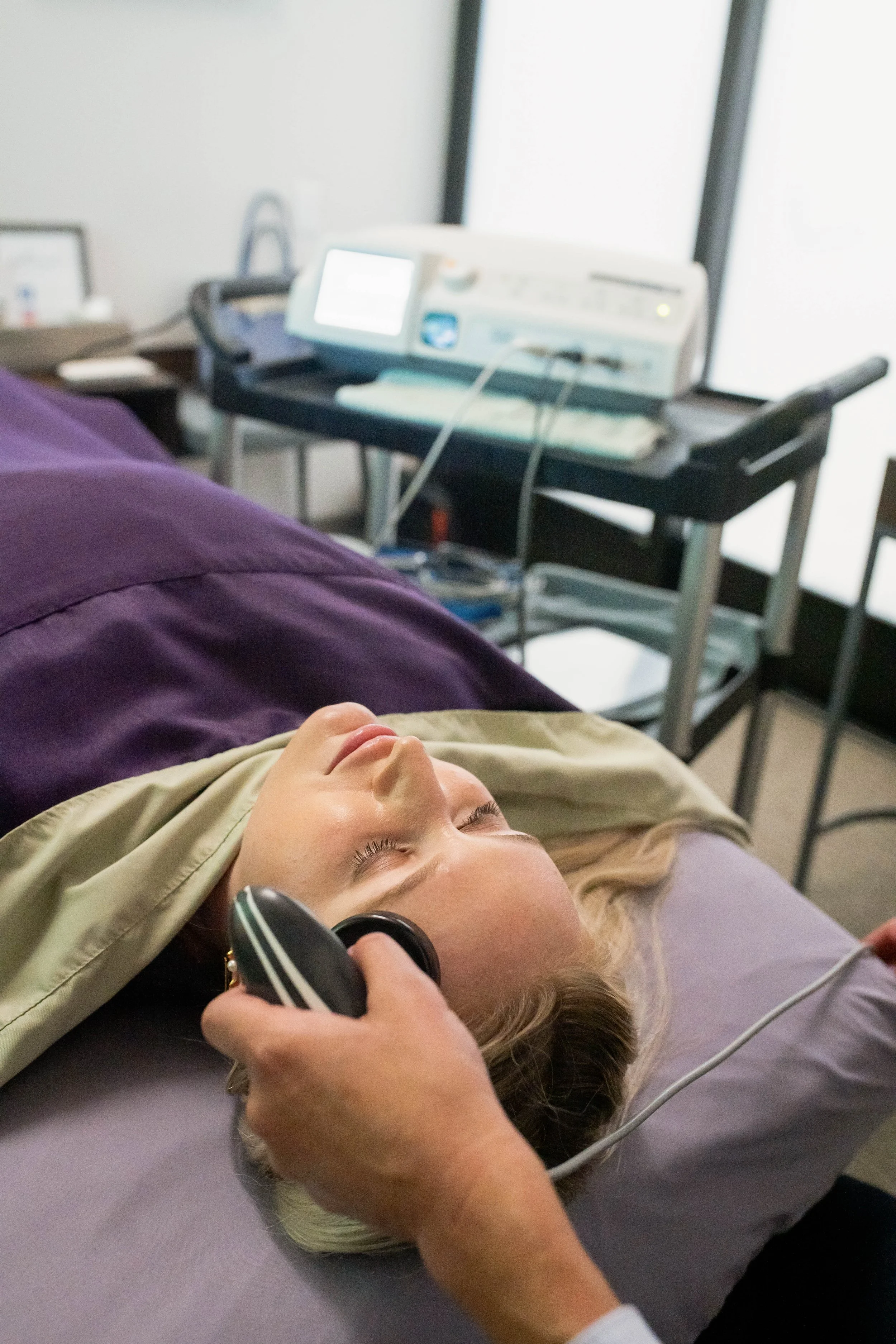 A person lying on a medical bed with their eyes closed, using headphones, with an EKG device attached to their chest, and a medical monitor and equipment in the background.
