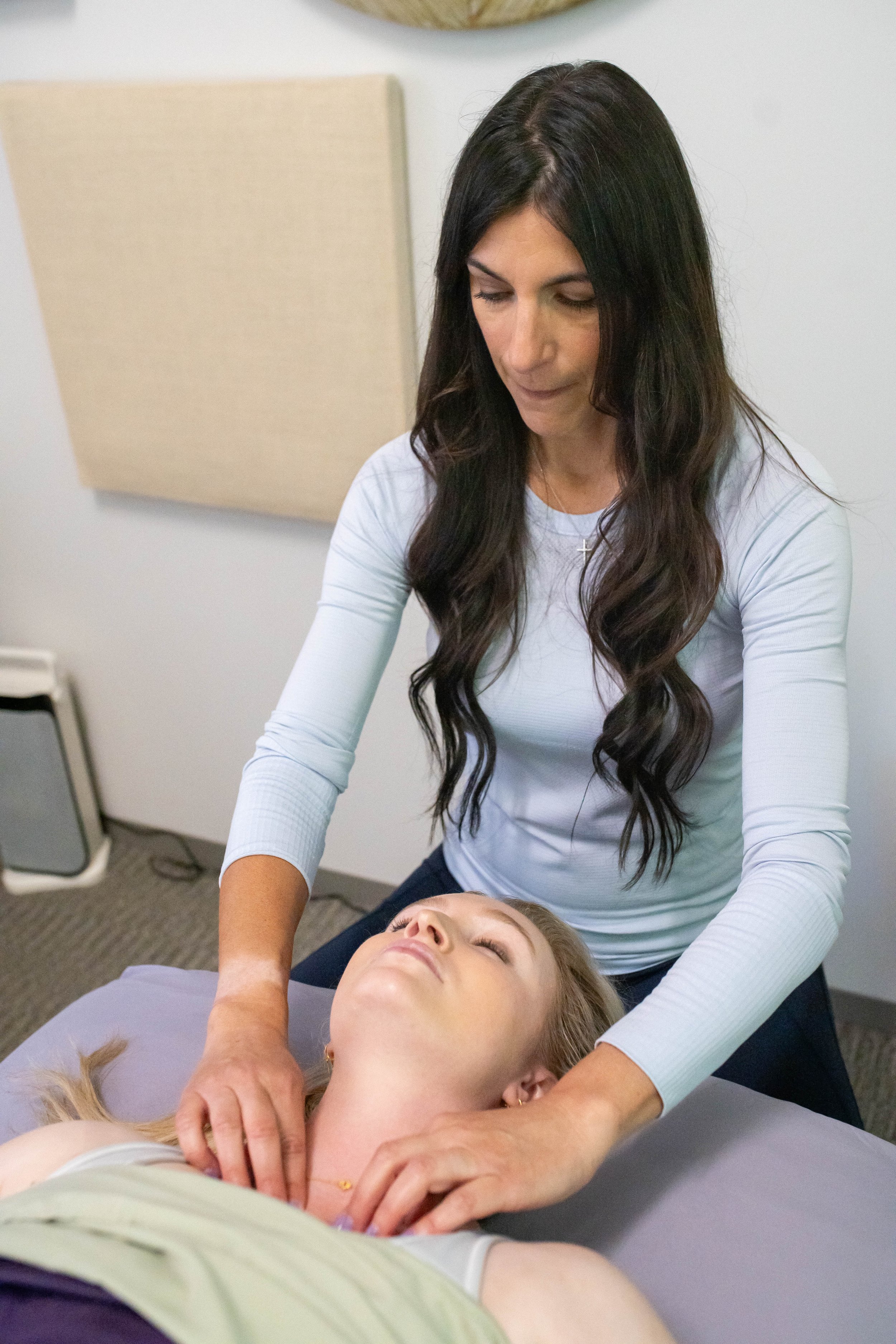 A woman with long dark hair giving a massage to another woman lying face up on a massage table, in a clinical setting.