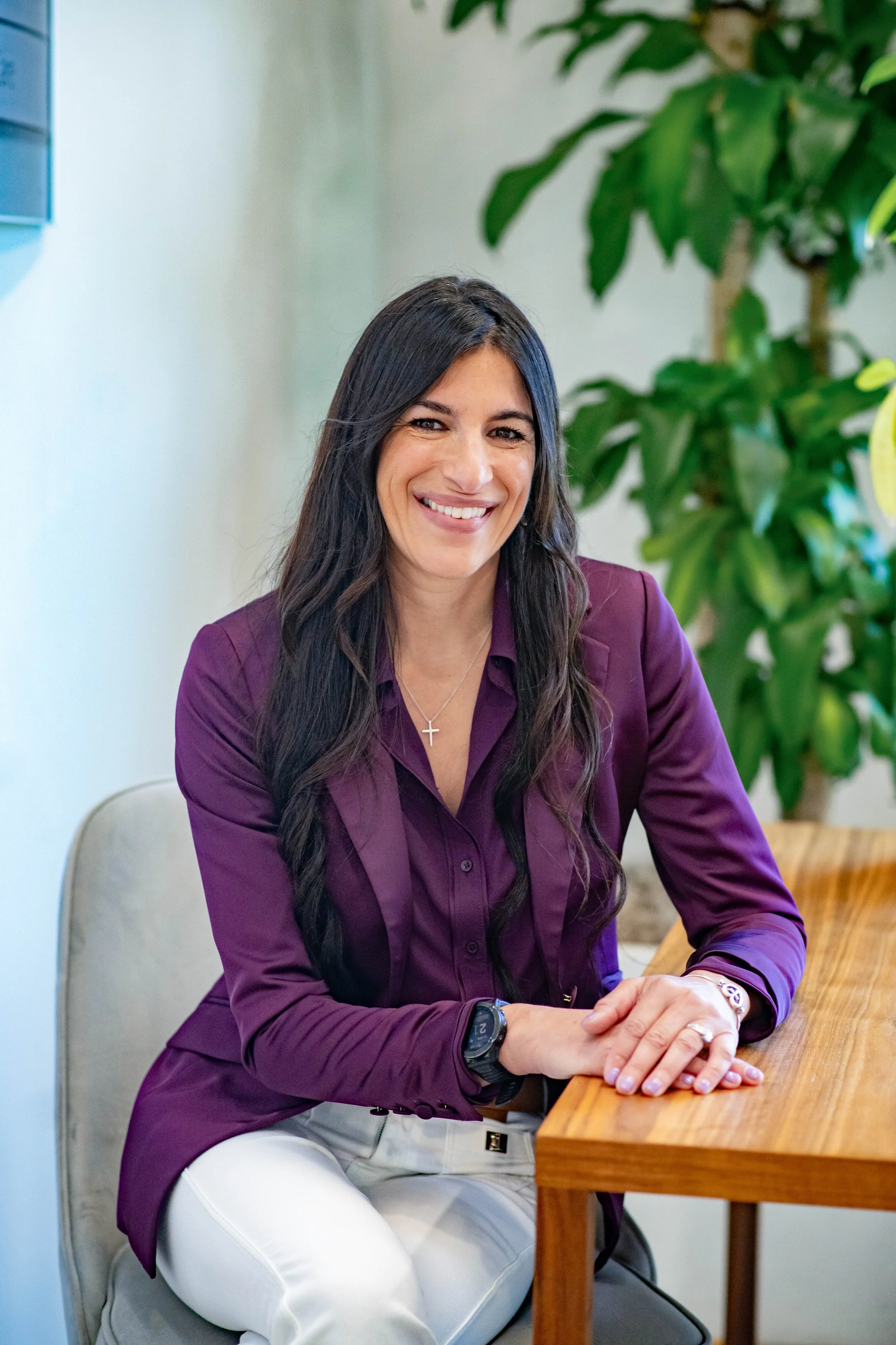 A woman with long dark hair smiling, sitting at a wooden table, wearing a purple blazer, white pants, and a watch, in a room with a large green plant in the background.