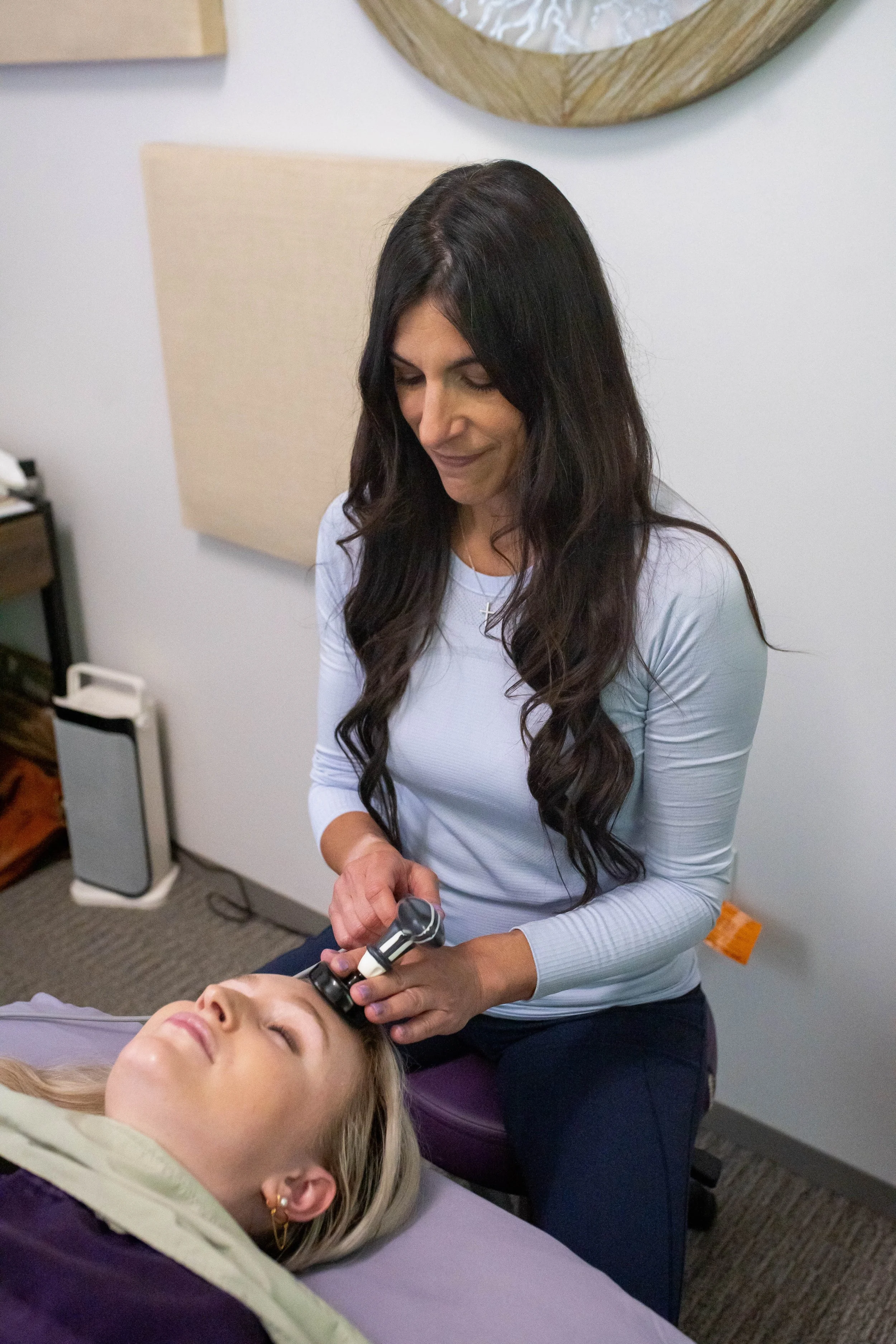 A woman with long dark hair receiving a facial treatment with a handheld device from another woman. The client is lying down with eyes closed, and the practitioner is standing beside her in a professional setting.