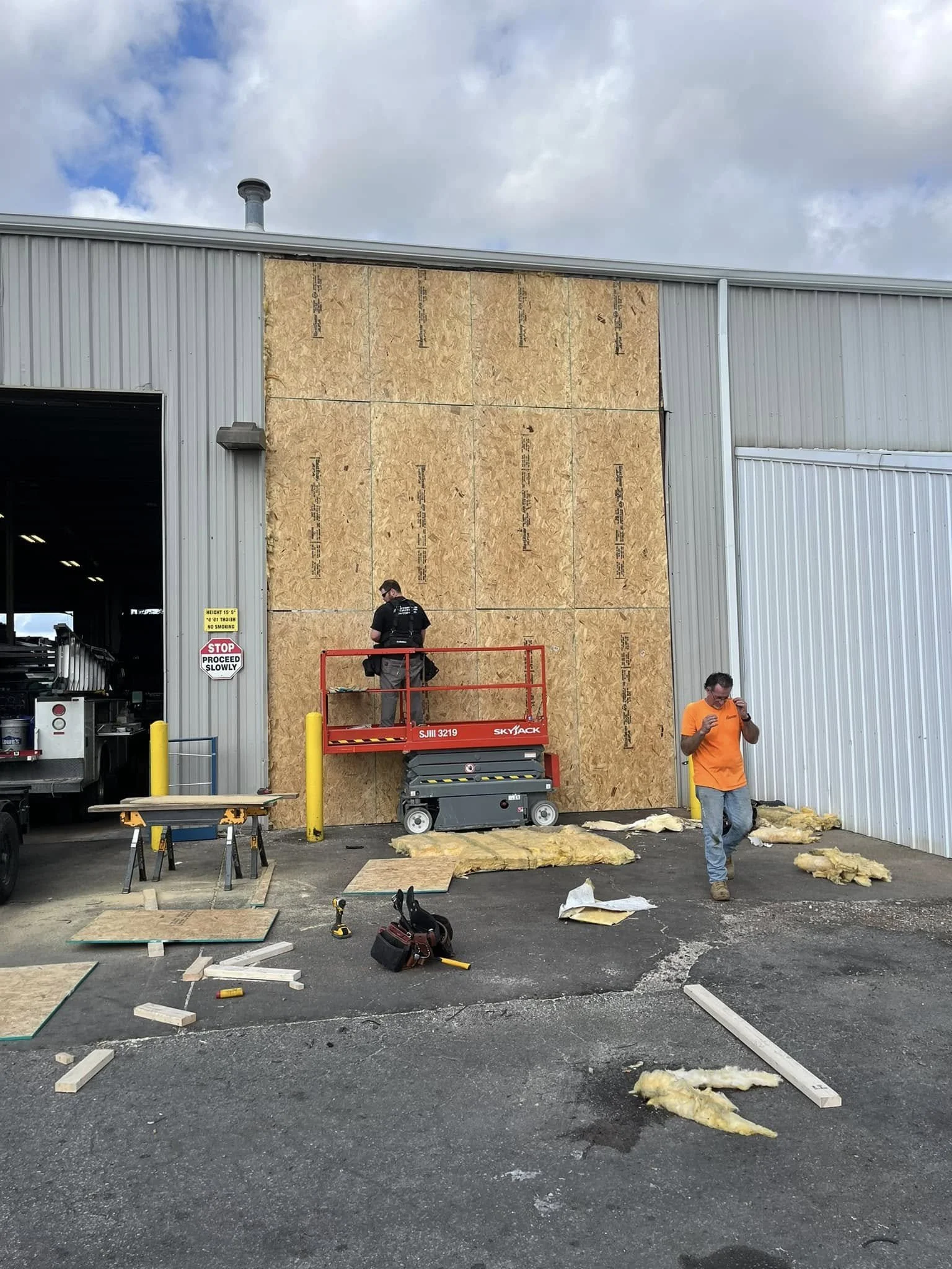 Construction workers are installing insulation on a building's exterior wall. One worker is on a lift, attaching sheathing, while another stands nearby, holding tools. Construction materials and tools are scattered on the ground.
