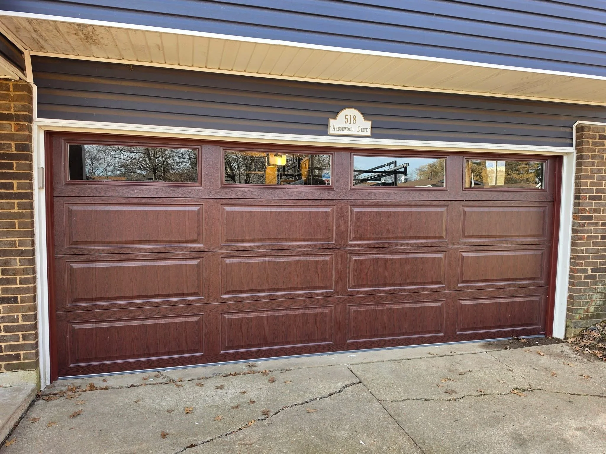 A brown garage door with small rectangular windows at the top, set in a brick wall. Above the door is a house number sign reading '518 Arborwood Drive'. The concrete driveway in front has some cracks and fallen leaves.