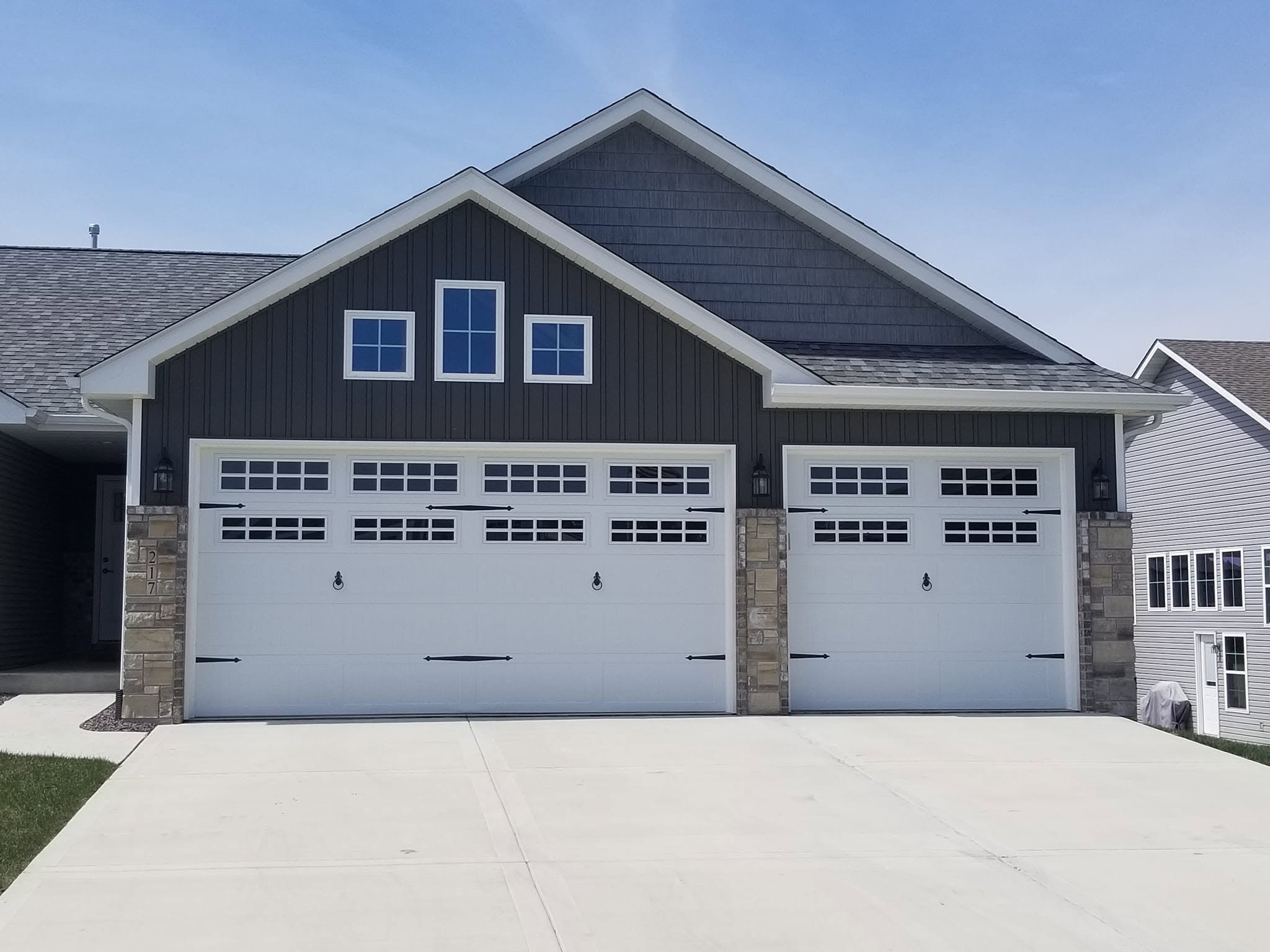 Front view of a house with a double garage door, dark gray siding, stone columns, and a triangular gable with three rectangular windows. A concrete driveway in front.
