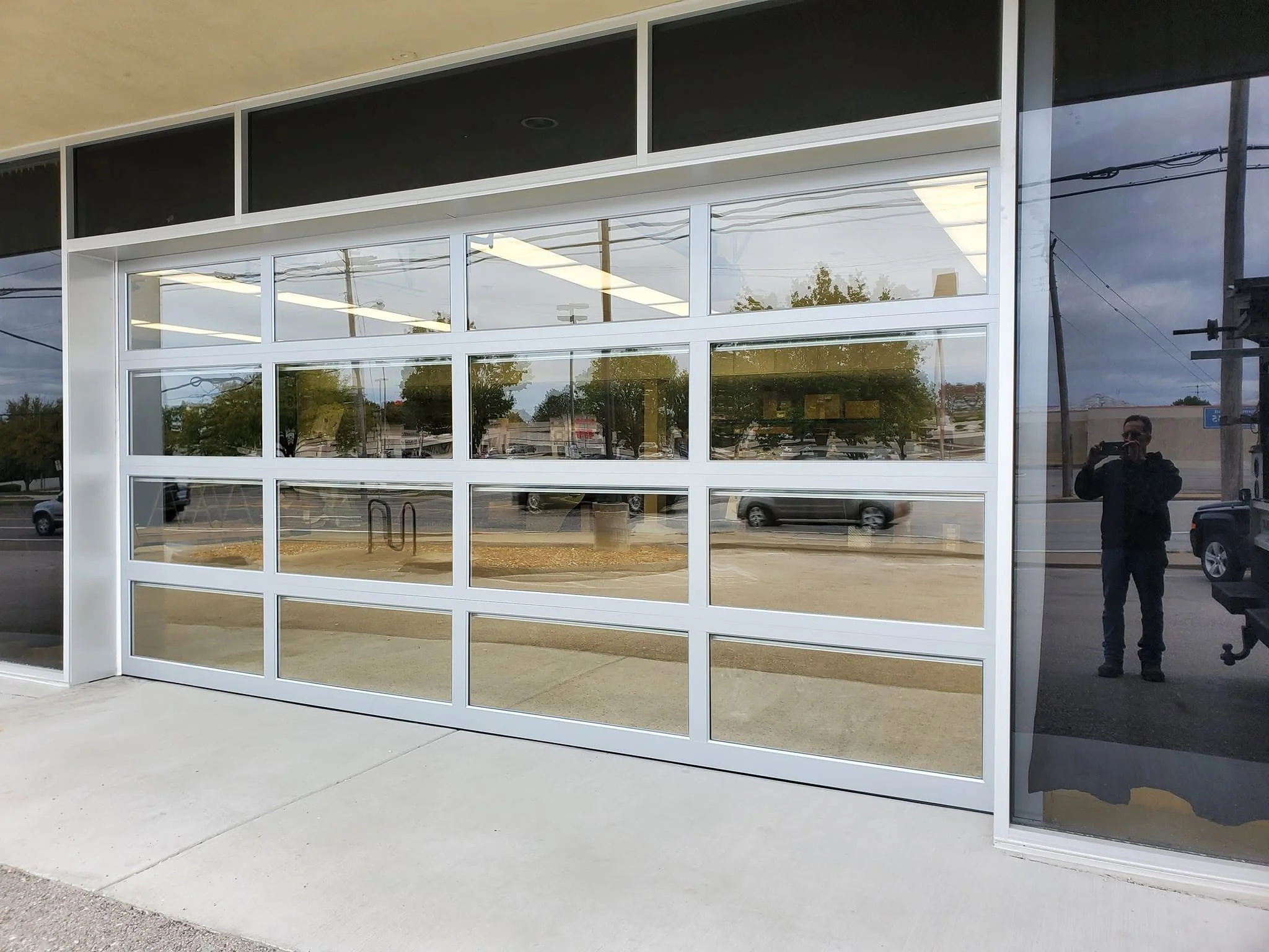A modern building with a large glass storefront. The exterior features white framing around the glass panels. The reflection in the glass shows parked cars, power lines, and a person taking the photo. The ground is made of concrete, and the sky appears cloudy.