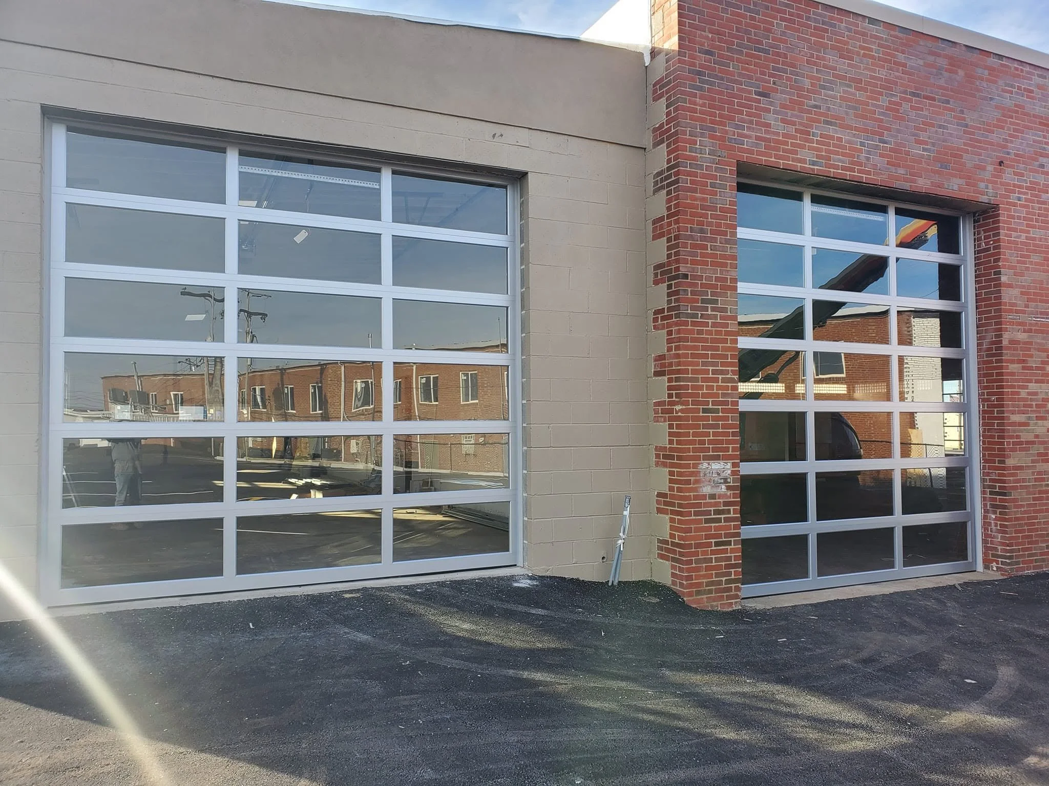 Exterior view of a building with large glass garage doors, beige and red brick walls, and a black asphalt ground.
