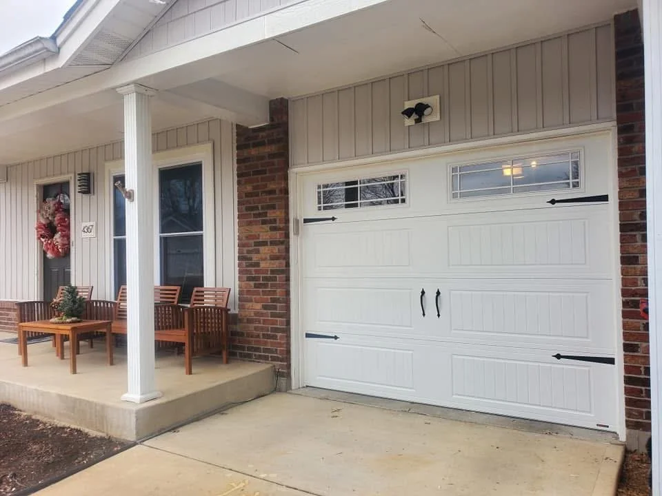 Front porch with Christmas wreath on the door, two wooden benches, a small table with a potted plant, and a white garage door with decorative hardware.