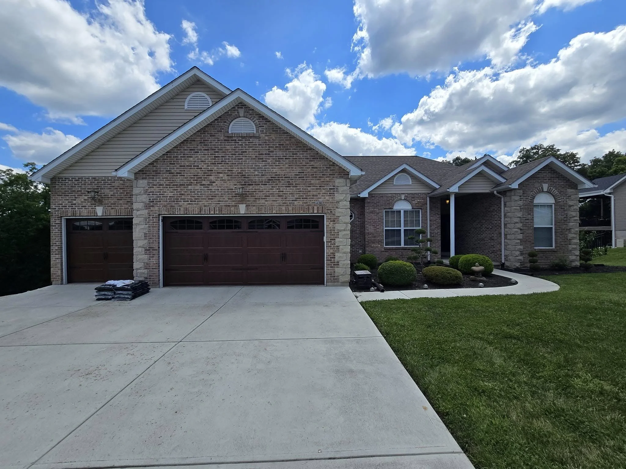 Front view of a brick house with a brown garage door, a driveway, and a well-maintained lawn with shrubs, under a partly cloudy blue sky.