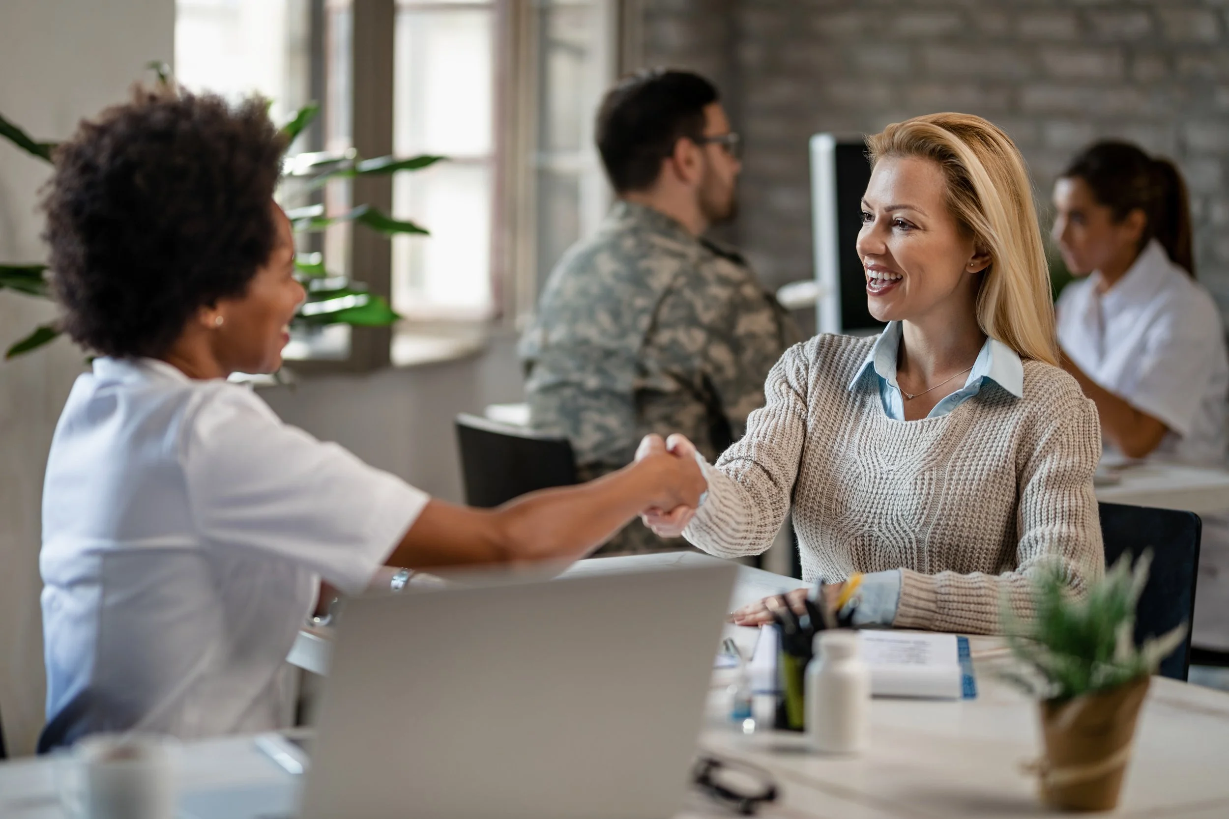 A woman with blonde hair and a beige sweater shaking hands with another woman in a white shirt in a meeting room, with other people sitting at a table, smiling and engaging in conversation.
