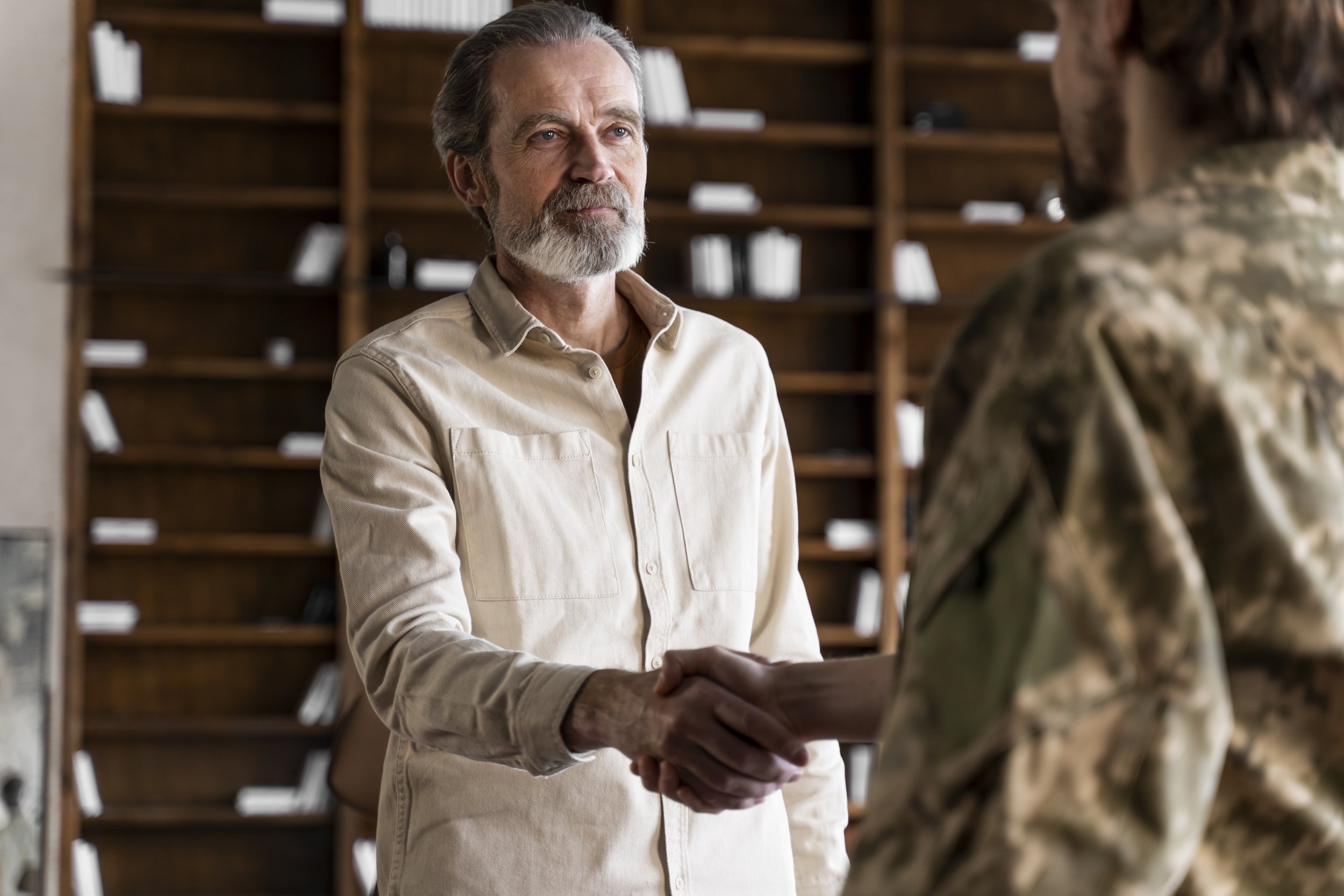 An older man with a beard and gray hair, wearing a beige shirt, is shaking hands with a person in military camouflage clothing inside a room with wooden shelves filled with white books or files.