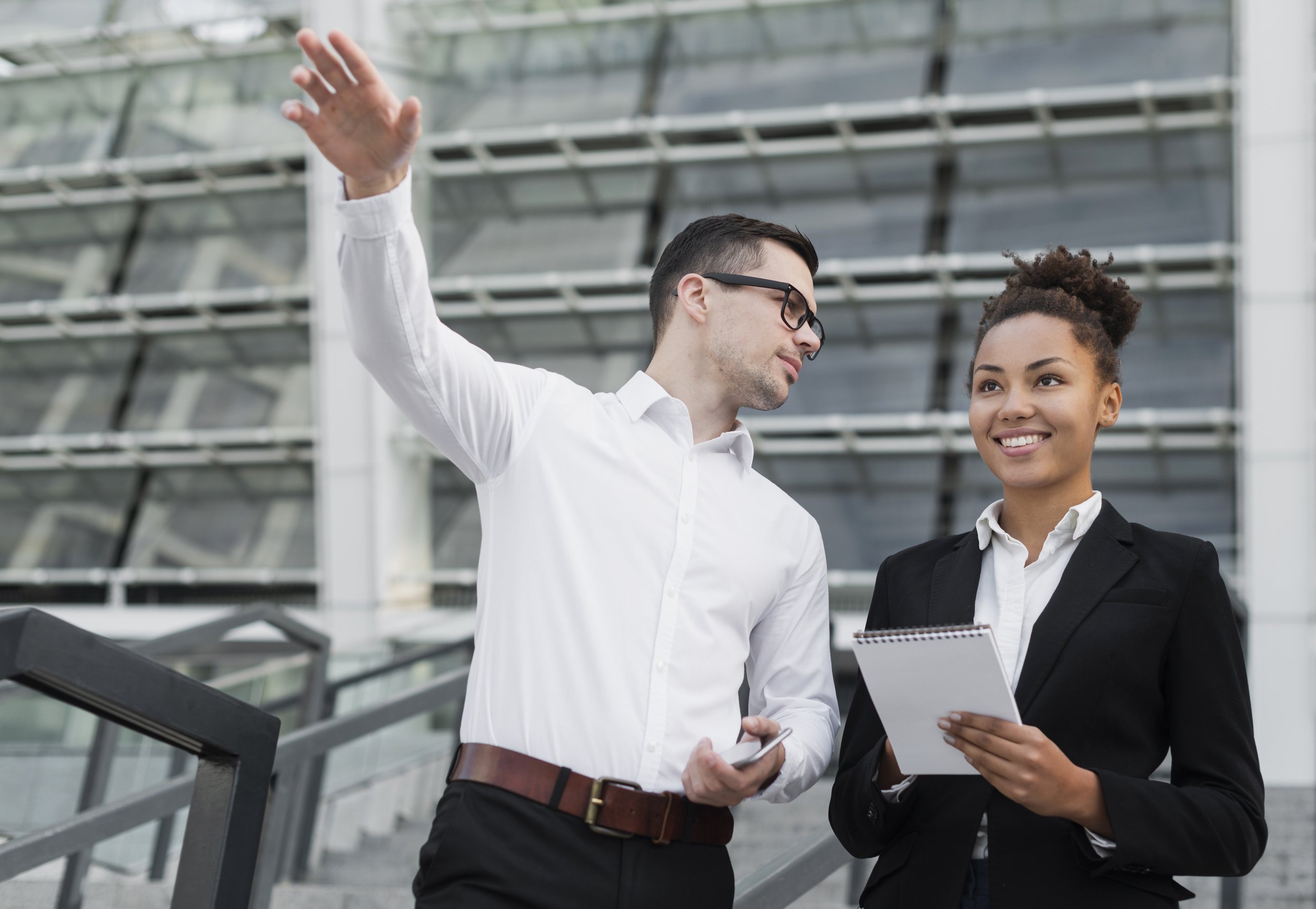 A man and woman dressed in business attire standing and talking inside a modern building with glass and metal architecture. The man is gesturing with his right hand while holding a phone in his left hand. The woman is holding a notebook and listening to the man, smiling.
