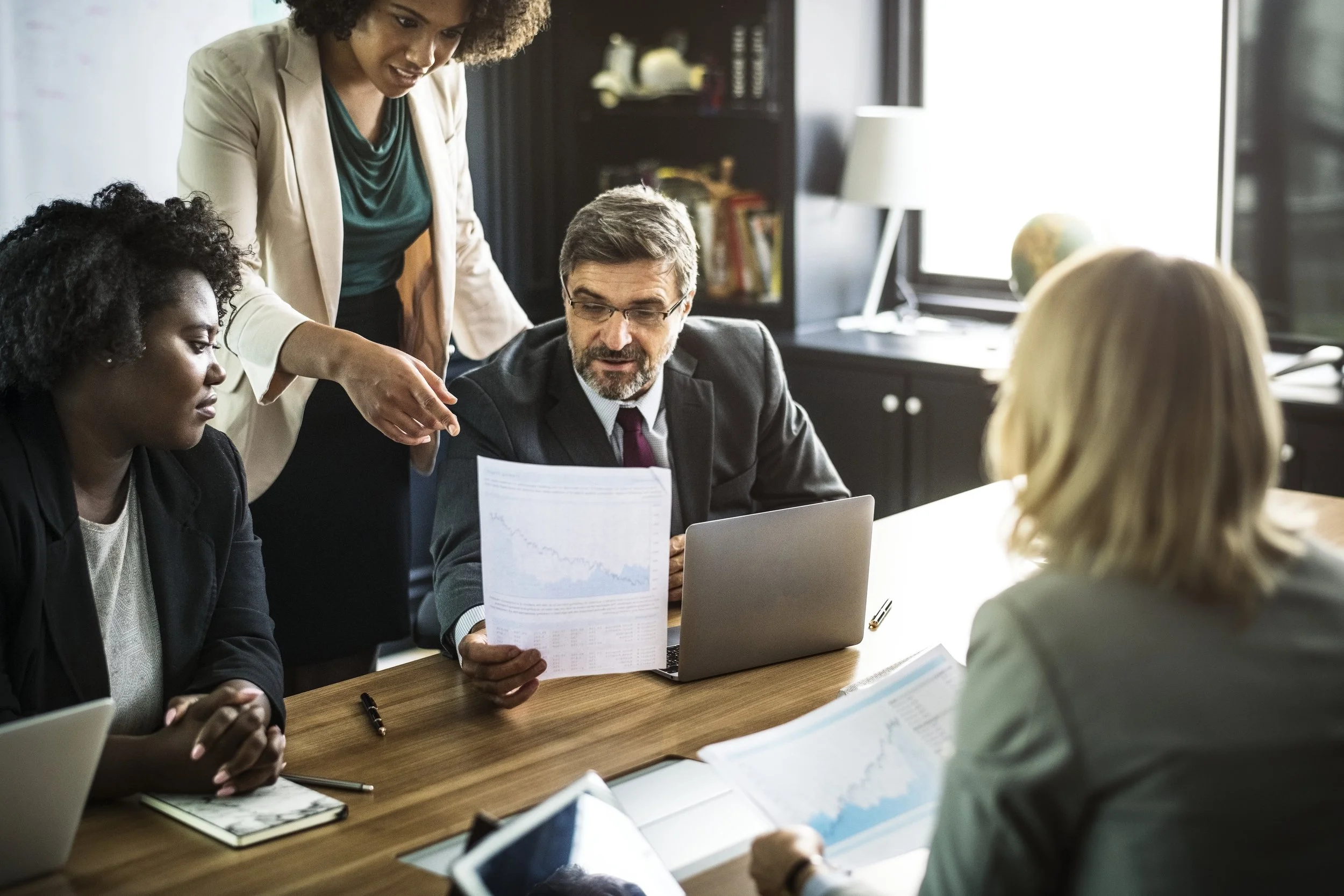 A diverse group of five business professionals gathered around a conference table discussing documents and a laptop in an office setting.