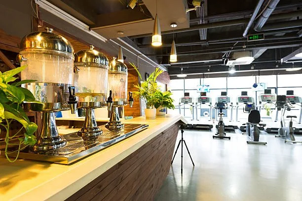 A modern gym with a breakfast station in the foreground and cardio equipment in the background.