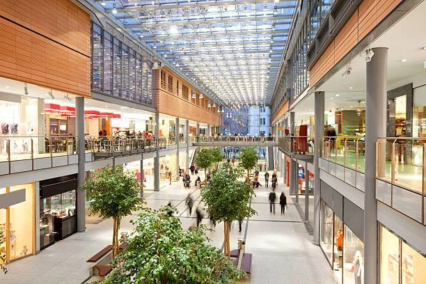 Interior view of a modern shopping mall with multiple levels, glass ceiling, trees, and shoppers walking.